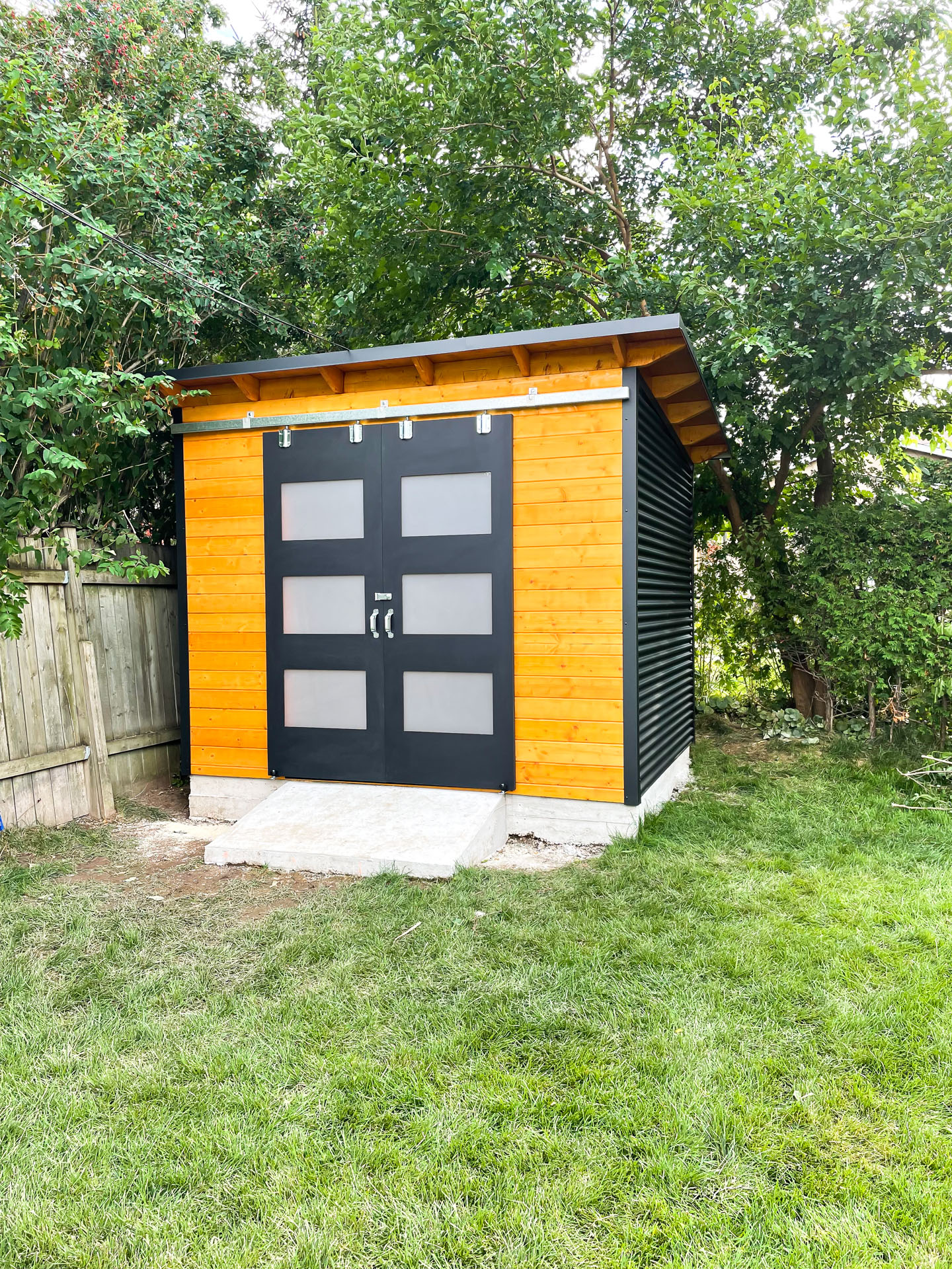 10x8 Essential Shed with jet black corrugated steel siding, Muskoka Brown stained pine front wall, and double sliding frosted glass barn doors in Burlington Ontario, installed by Backyard Escape Studios.