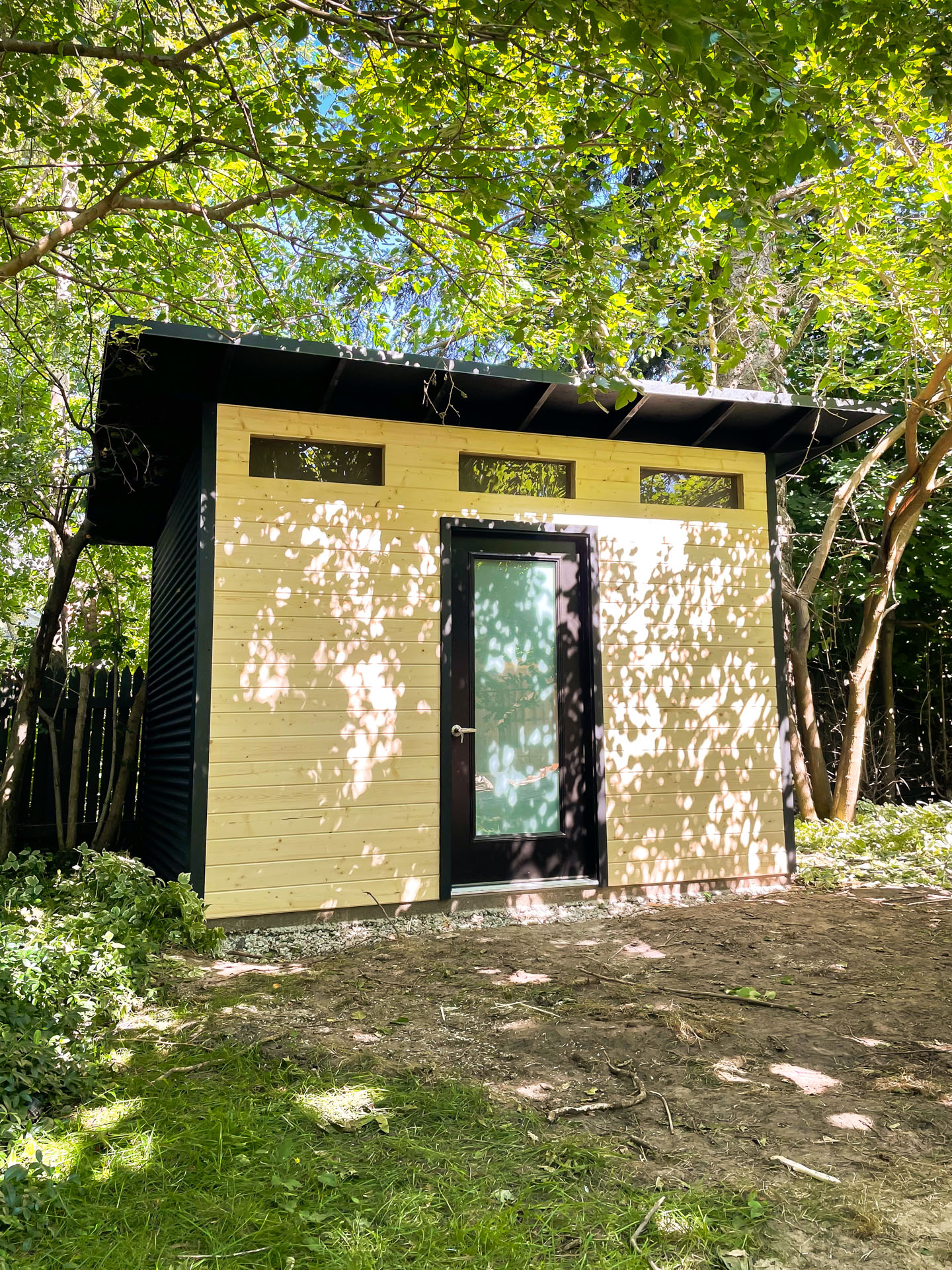 12x9 modern shed with jet black corrugated steel siding, clear-coat pine front wall, and frosted glass inswing door installed among trees in Blue Mountains Ontario by Backyard Escape Studios.