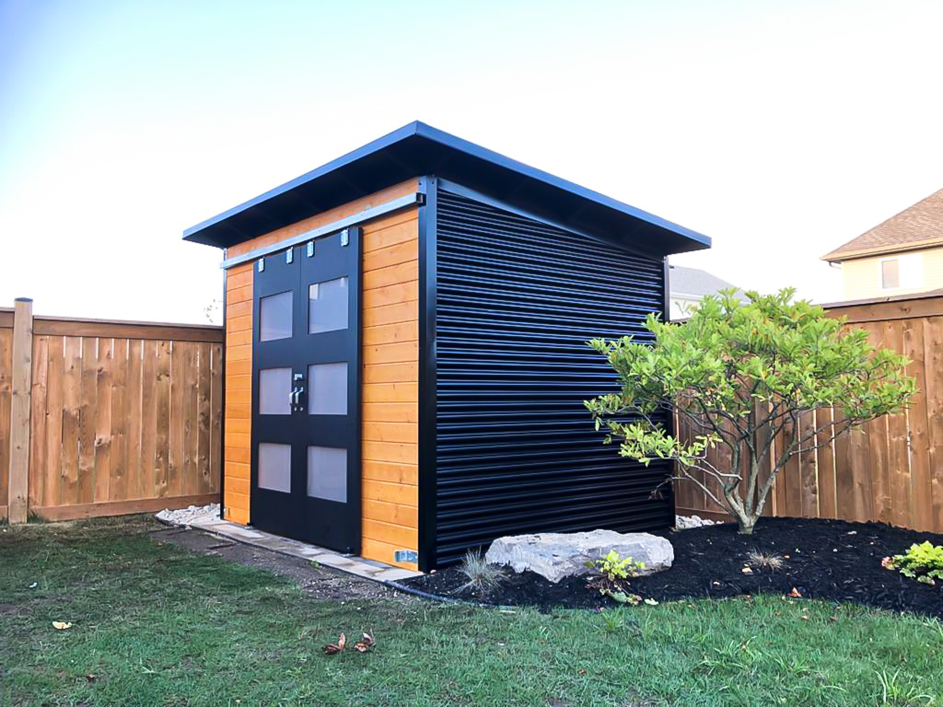 10x8 Essential Shed with cedar stain pine front wall and black double sliding barn doors with frosted glass, installed in a St. Thomas Ontario backyard by Backyard Escape Studios.