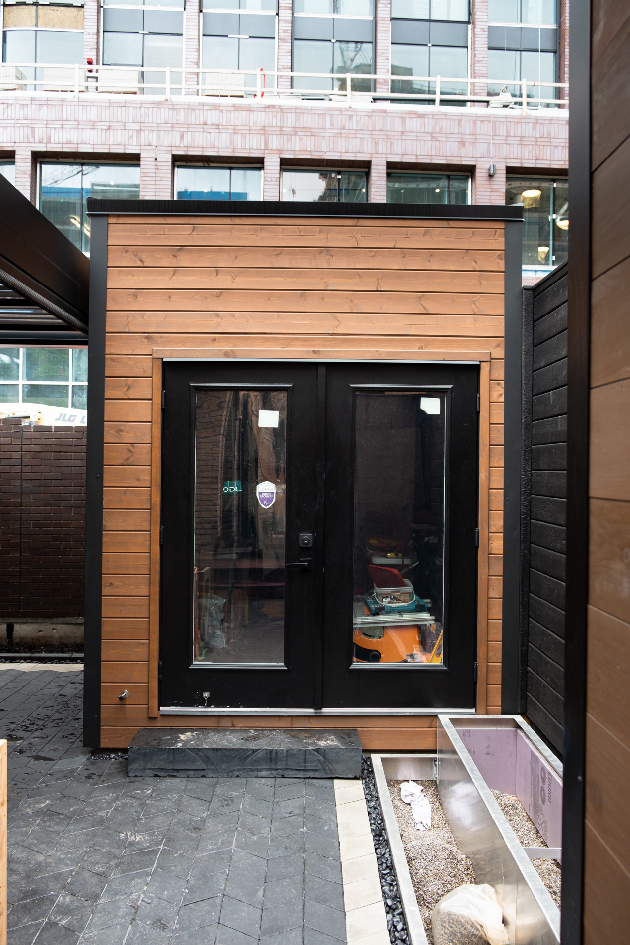 8x9.5 Cube Shed backyard studio with cedar tongue-and-groove front wall in Muskoka Brown stain, Jet Black corrugated steel siding, and black French doors with clear glass panels in Toronto Ontario, installed by Backyard Escape Studios.