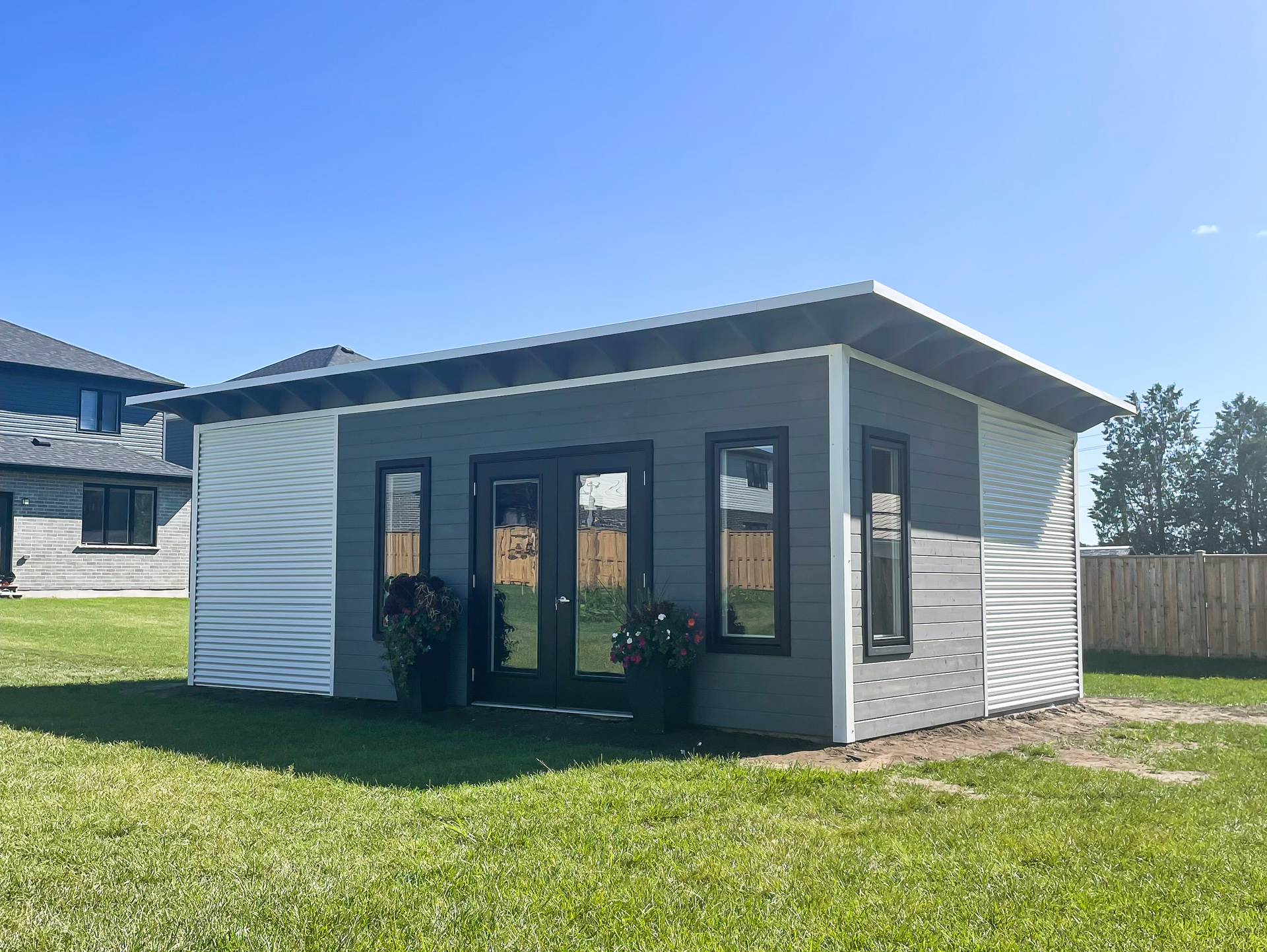 25x15 Advanced Shed backyard studio with Cambridge White corrugated steel siding, Kendall Charcoal stained pine front wall, full-glass French doors and roll-up garage door in Mount Brydges Ontario, installed by Backyard Escape Studios.