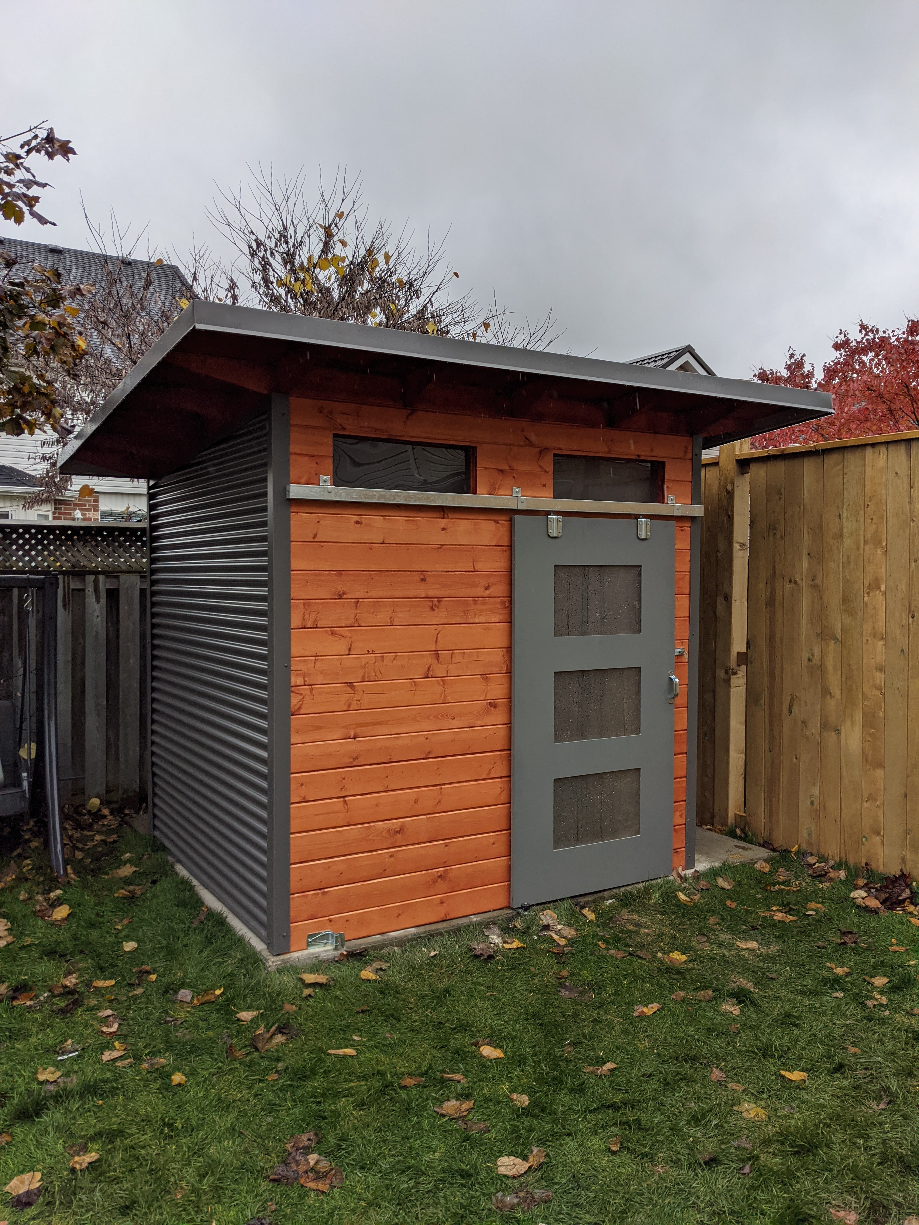 8x7 Advanced Shed with charcoal corrugated steel siding, mahogany-stained pine tongue-and-groove front wall, and sliding barn door with frosted glass panels in Guelph Ontario, installed by Backyard Escape Studios.