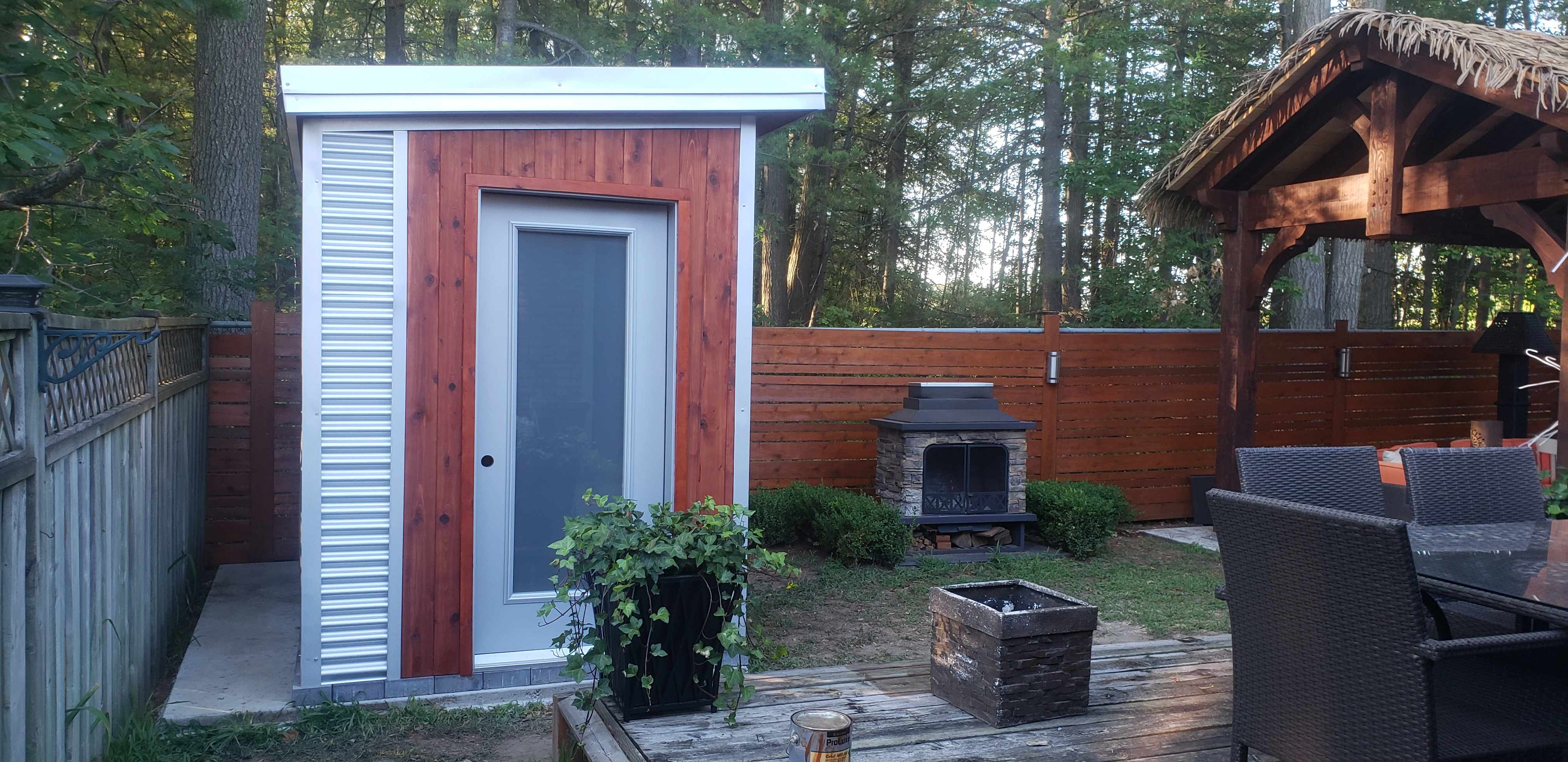 9x6 sauna shed with full-lite glass panel door set in backyard outdoor living area with stone fireplace in Barrie Ontario, installed by Backyard Escape Studios.