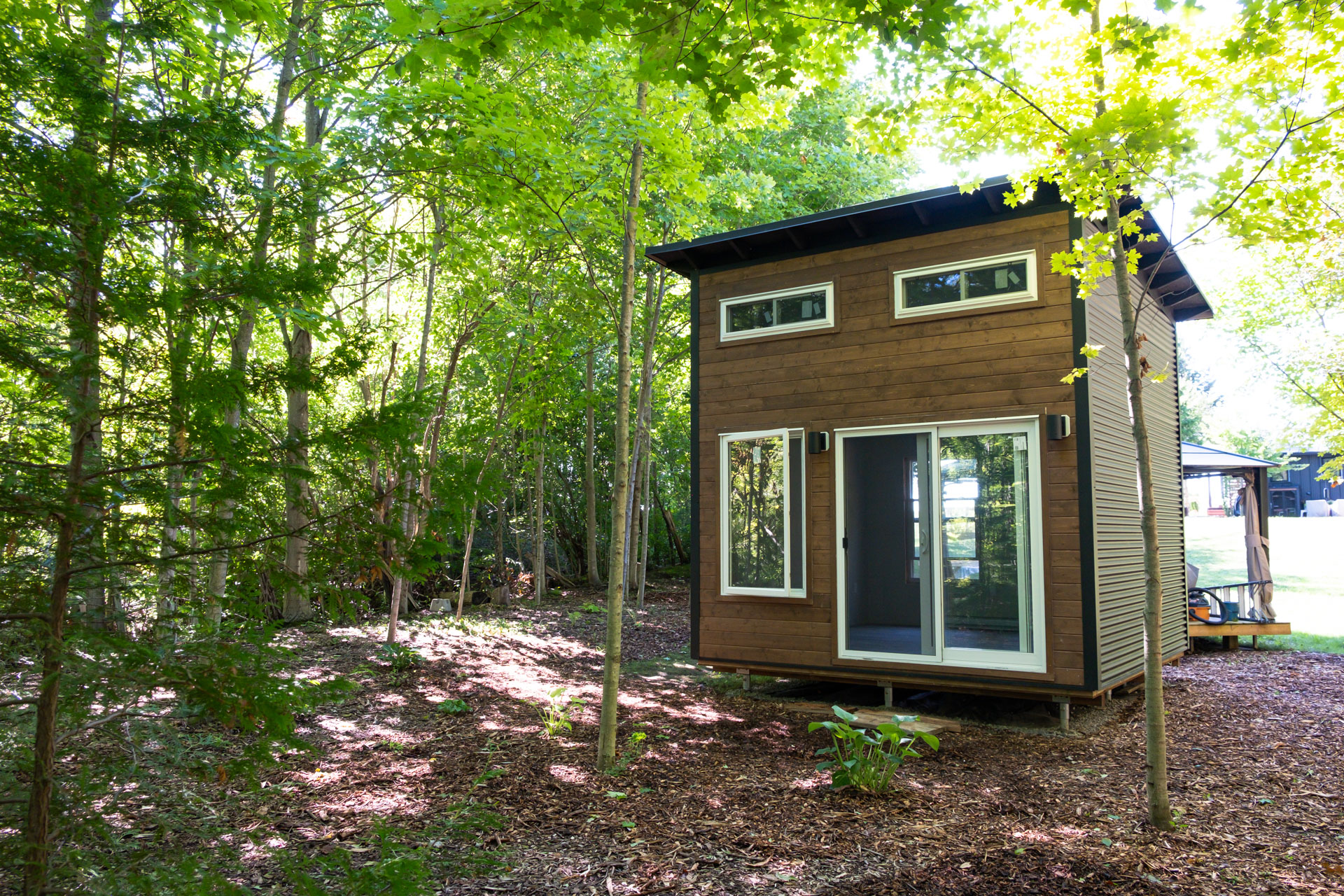 12x12 Essential Studio bunkie with Muskoka Brown pine T+G siding, Jet Black corrugated steel sidewalls, mono-slope black roof, and white patio sliding doors among trees in Bayfield Ontario, installed by Backyard Escape Studios.