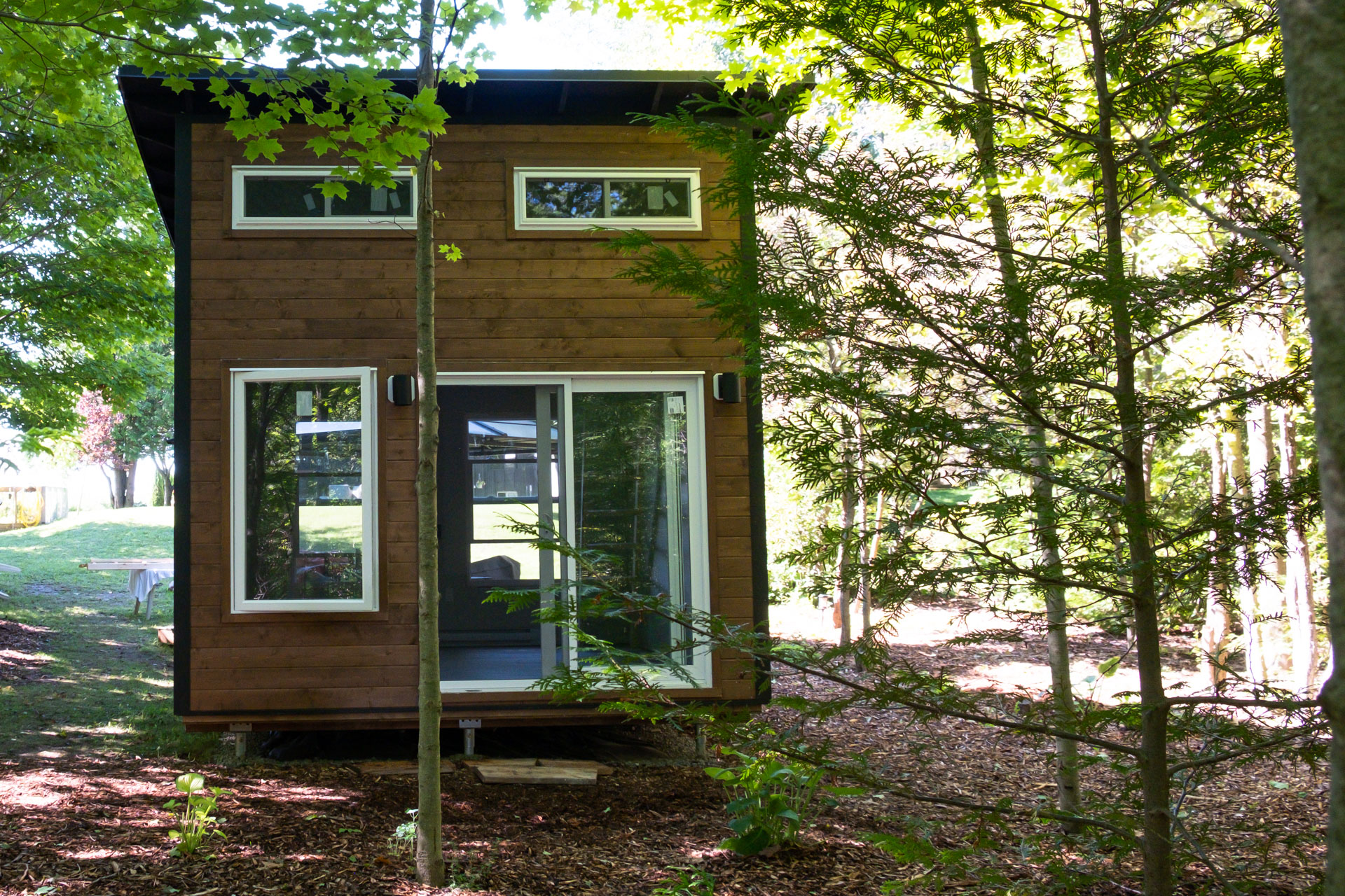 Side exterior view of 12x12 Essential Studio bunkie in Bayfield Ontario showing Jet Black corrugated steel siding, Muskoka Brown pine front wall, exposed rafter soffits, and white patio sliding doors on a wooded Huron County property, installed by Backyard