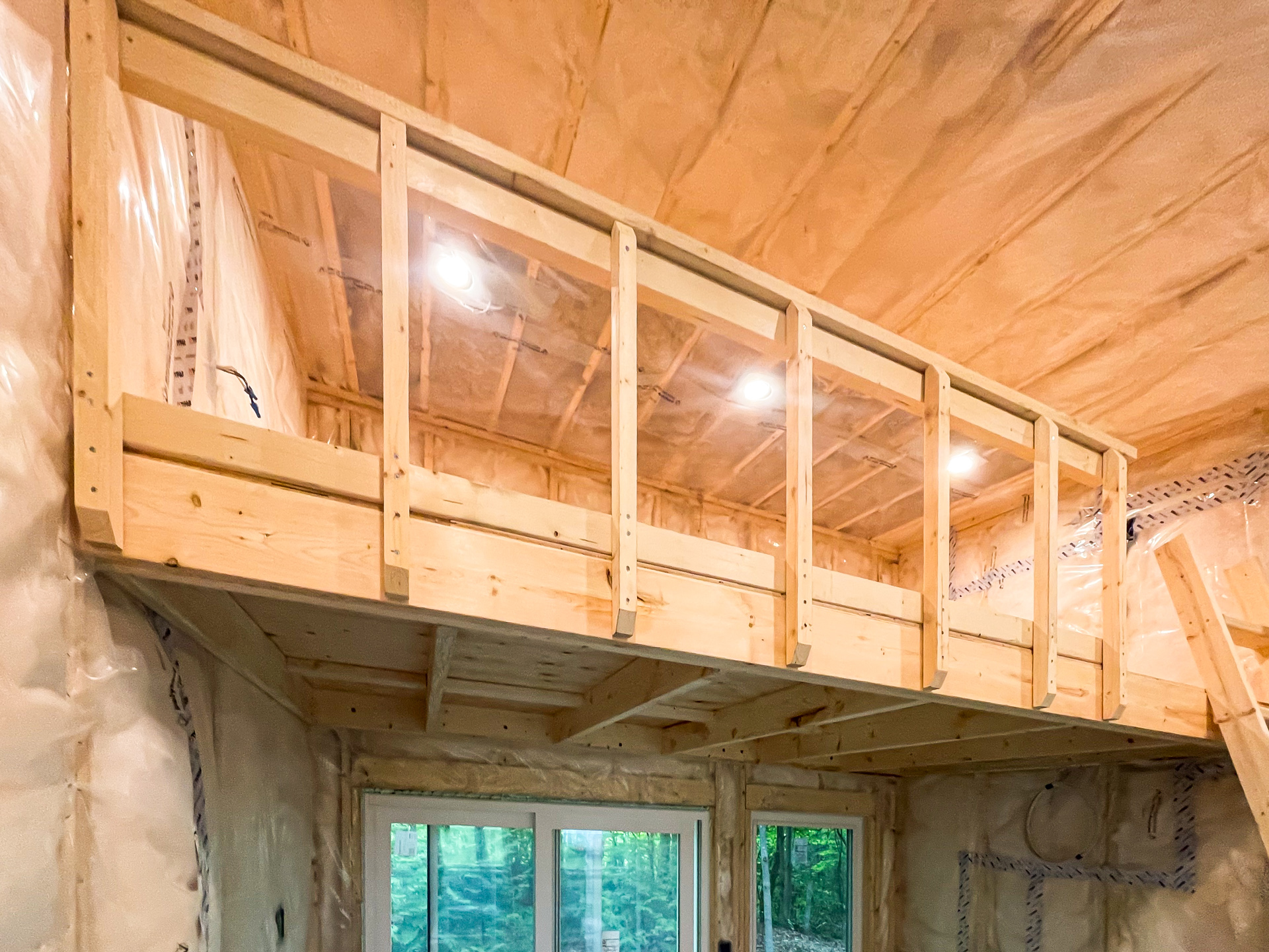 Interior view of 12x12 Essential Studio bunkie in Bayfield Ontario showing pine-framed sleeping loft with braided steel cable support, recessed lighting, vapour barrier walls, and white patio sliding doors below, installed by Backyard Escape Studios.
