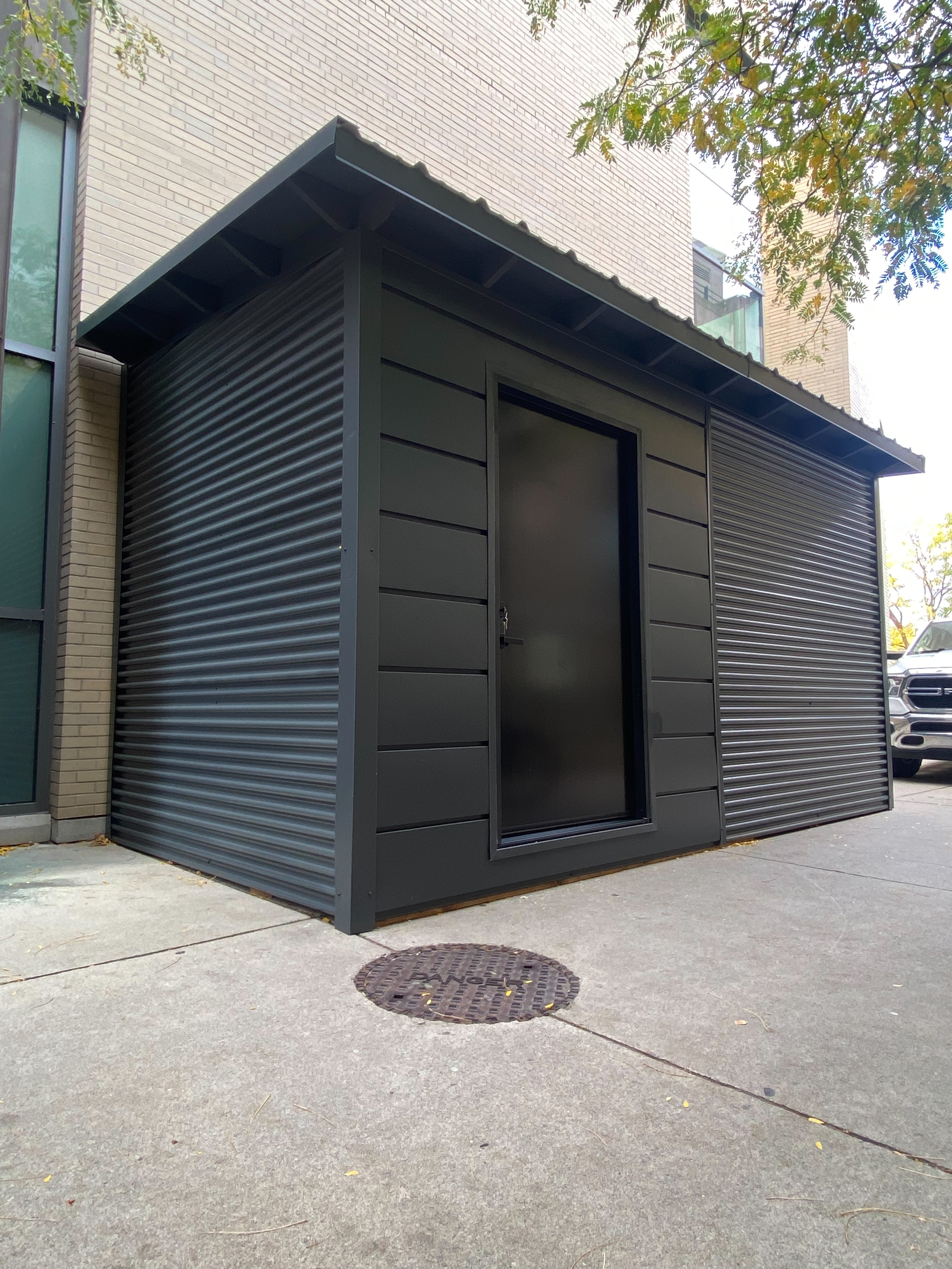 Corner angle view of 16x10 Essential Shed showing charcoal corrugated steel siding on side and front walls with steel roof overhang in Hamilton Ontario installed by Backyard Escape Studios.