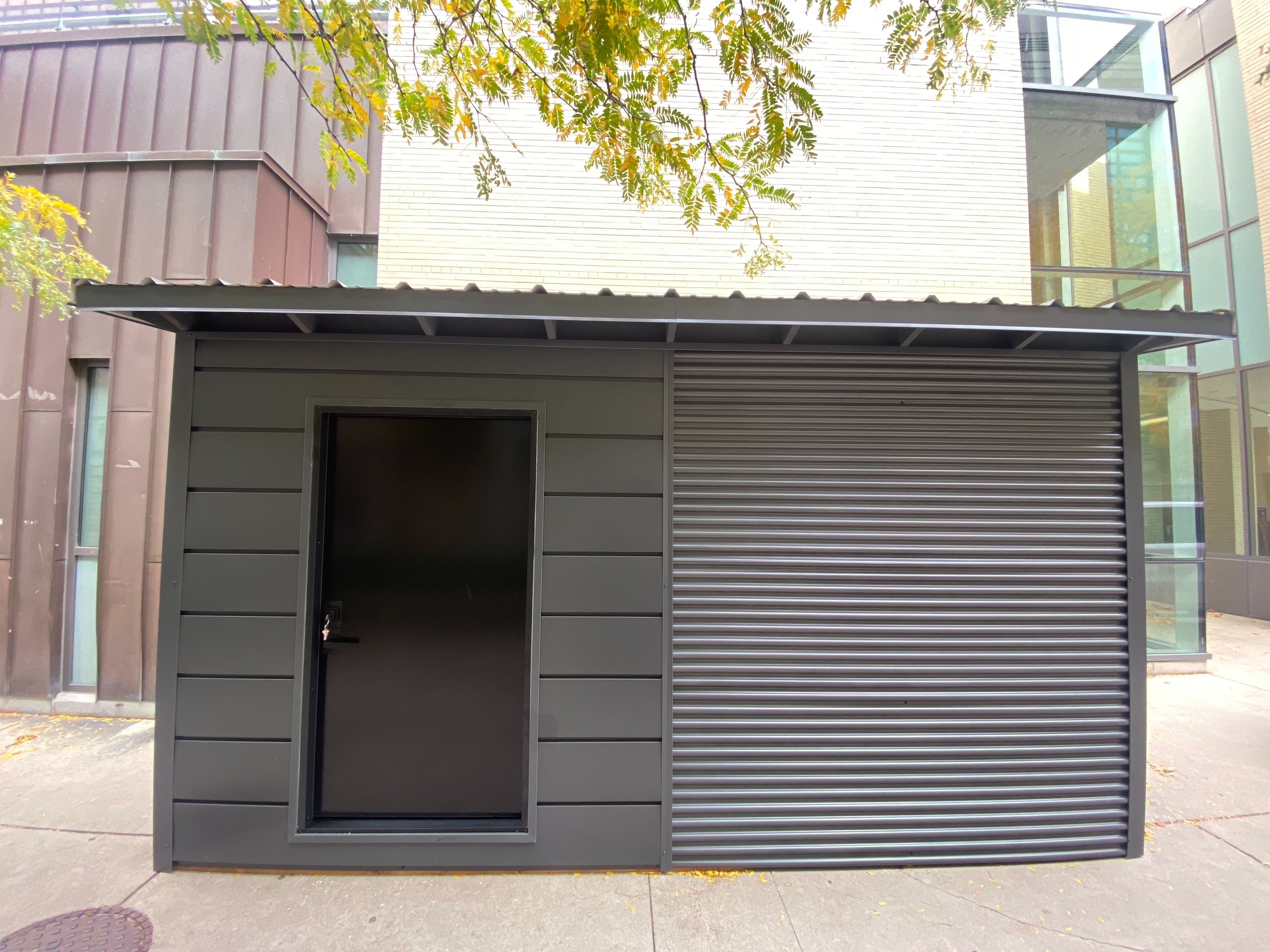 Straight-on front elevation of 16x10 Essential Shed with charcoal steel corrugated siding, flush door, and steel roof overhang in Hamilton Ontario installed by Backyard Escape Studios.