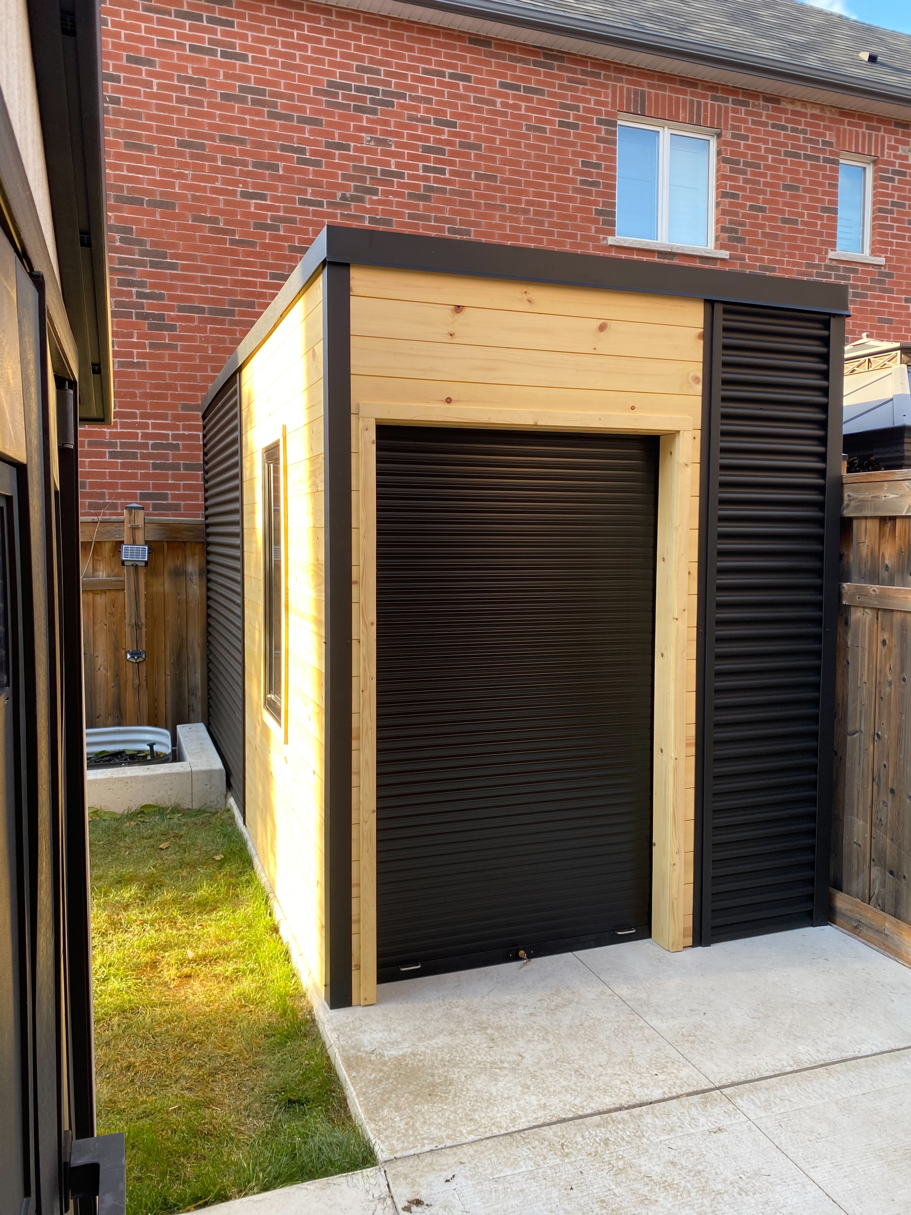 14x7 Modern Utility Shed with Ebony corrugated steel siding, natural pine T&G panelling, and aluminum roll-up shutter door in Mississauga Ontario, installed by Backyard Escape Studios.