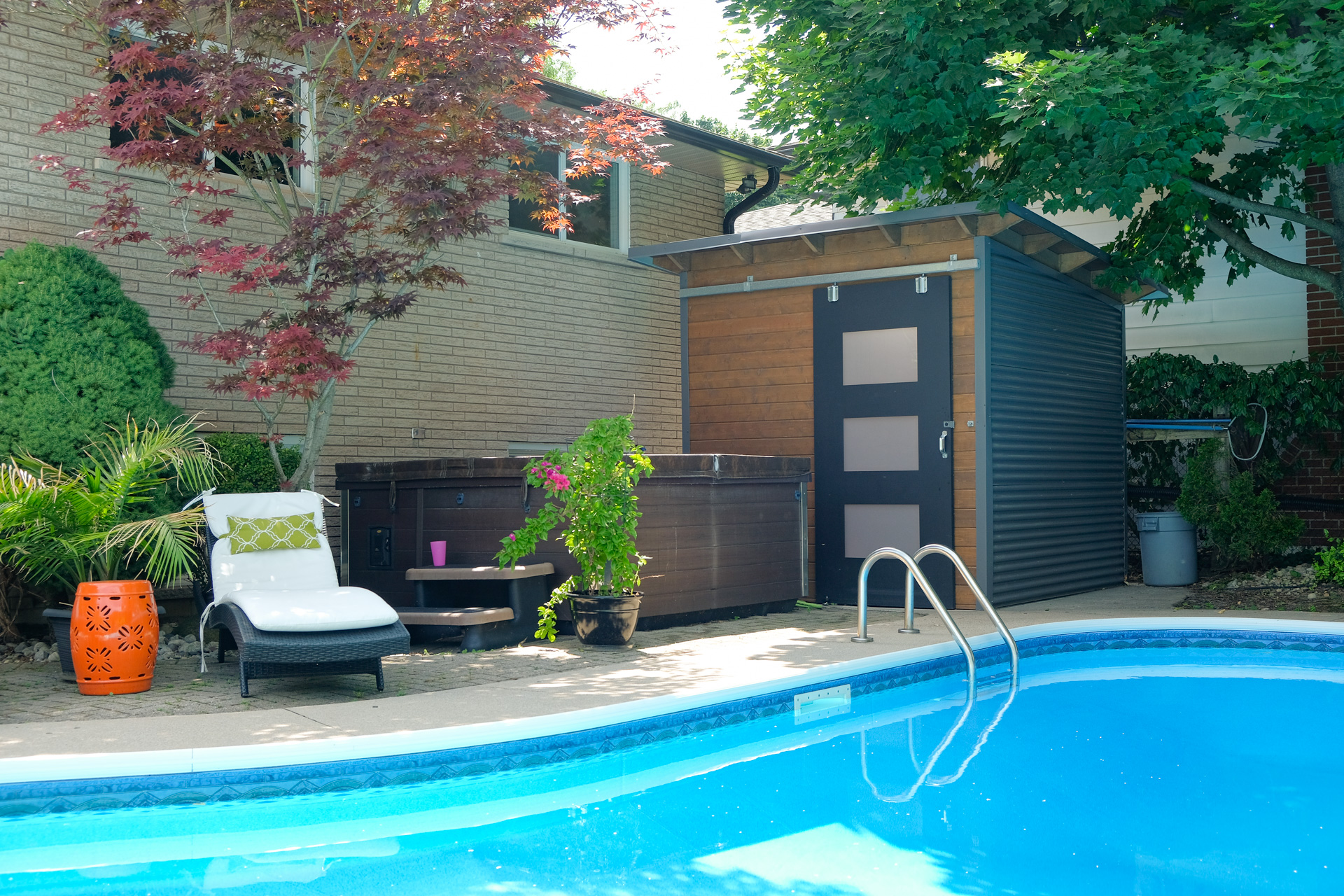 8x8 Essential Pool Shed with charcoal corrugated steel siding, Muskoka Brown pine front wall, and sliding barn door with frosted glass windows poolside in Hamilton Ontario, installed by Backyard Escape Studios.