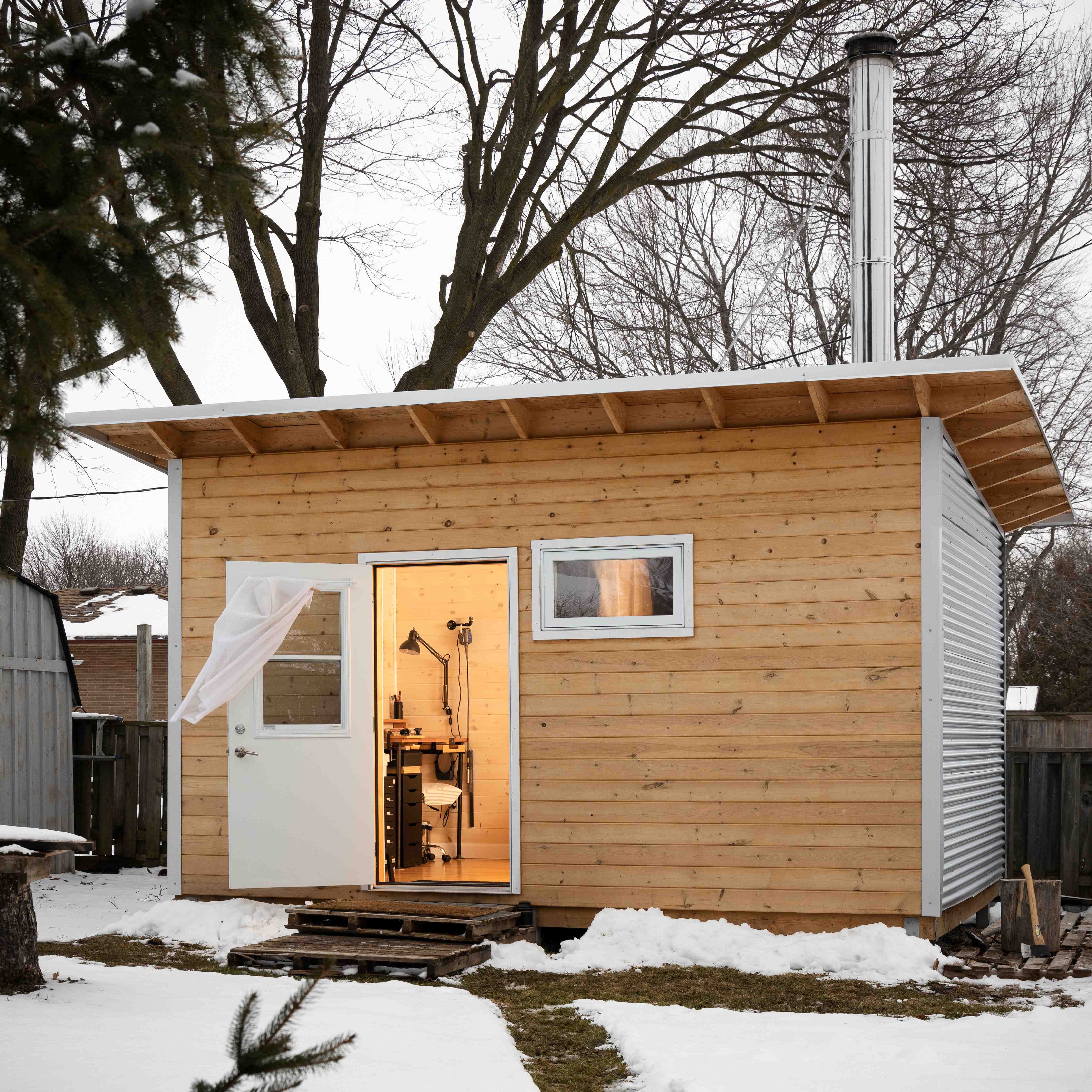 Interior of a backyard silversmith studio with black wood stove, pine tongue and groove walls, navy curtains, and laminate flooring in Ontario