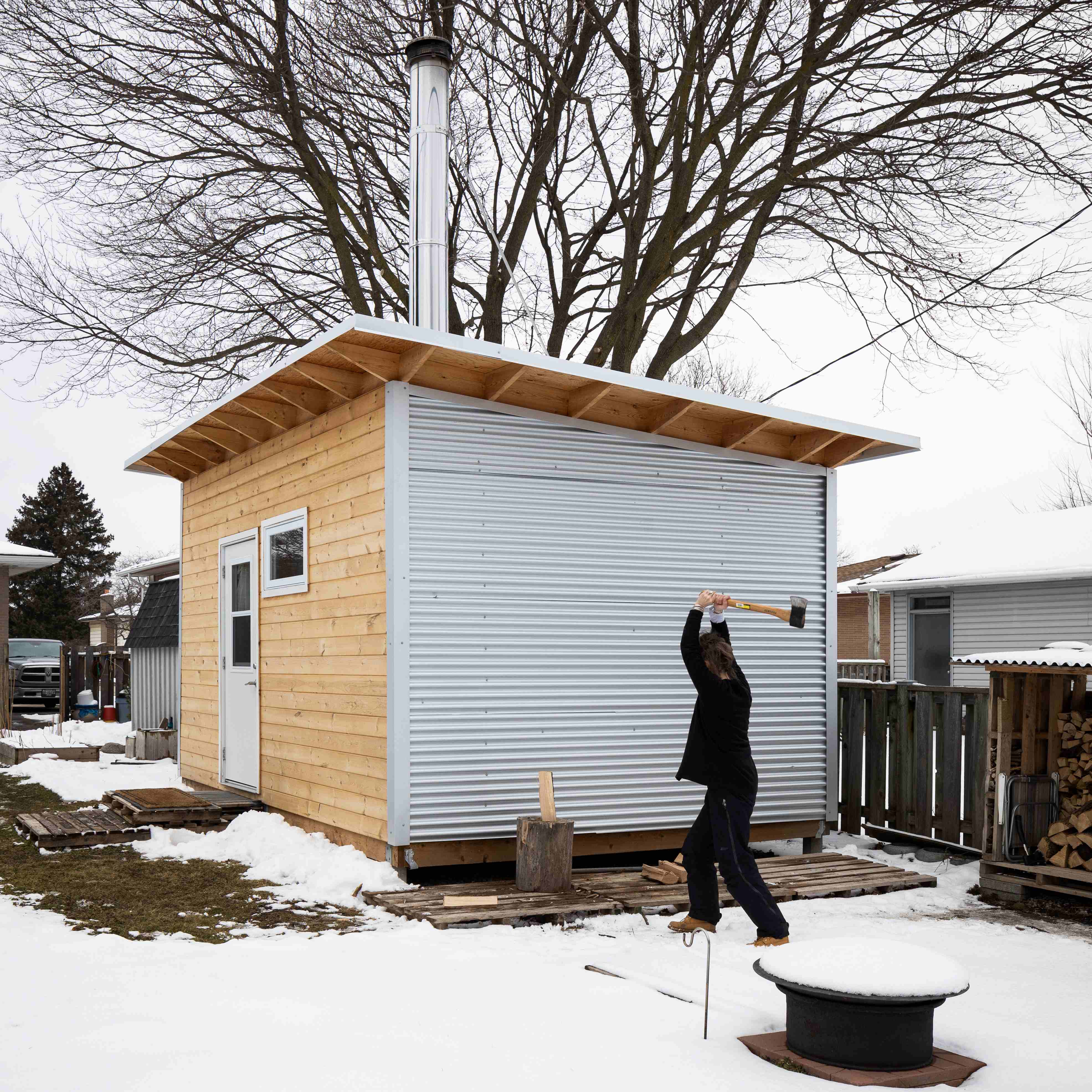 Side view of a backyard studio with Galvalume corrugated steel siding and pine front face, chimney pipe, and person splitting wood in a snowy Ontario backyard