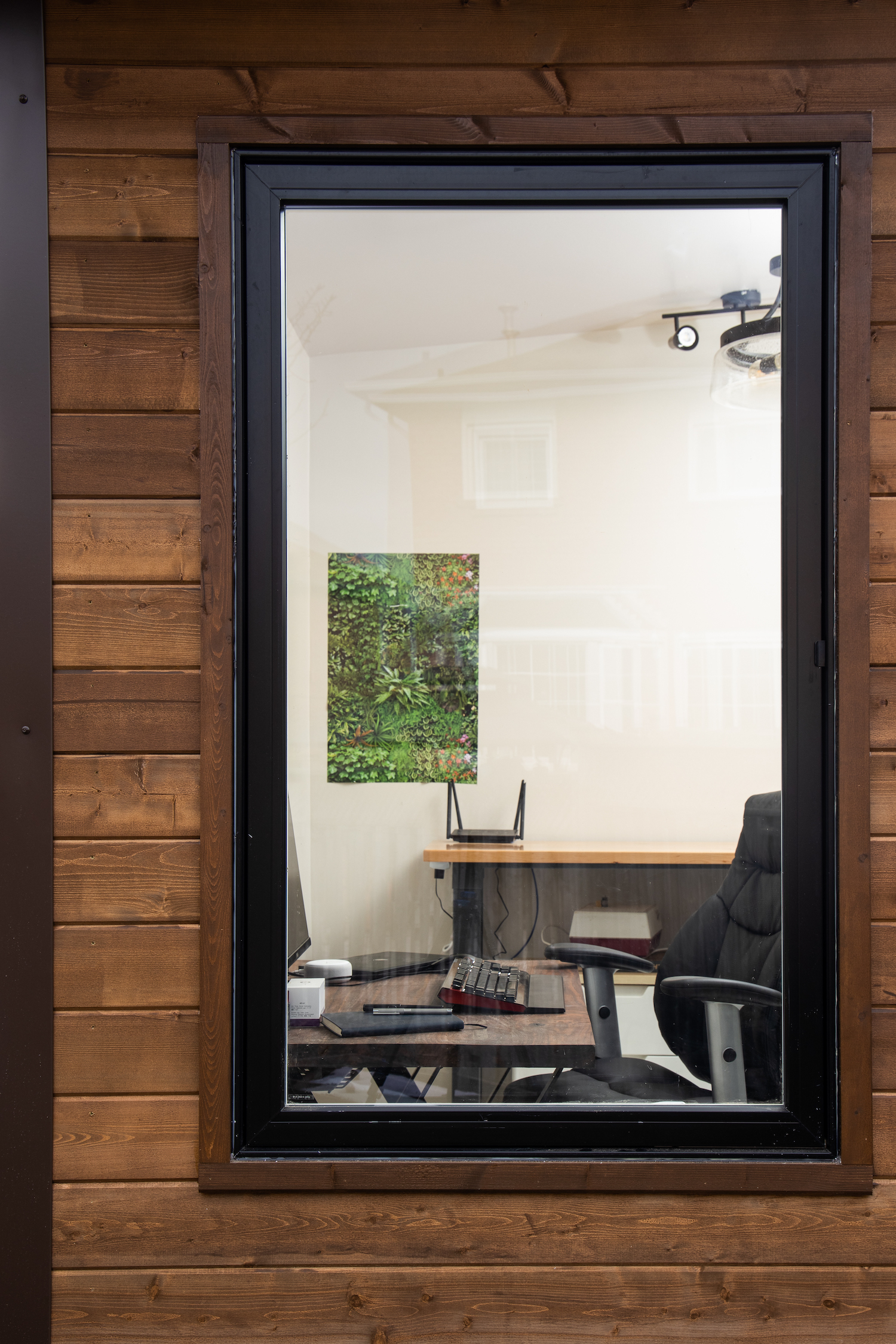 Large black-framed window view into a backyard home office showing desk setup and botanical art inside, Muskoka Brown pine siding in Mississauga Ontario