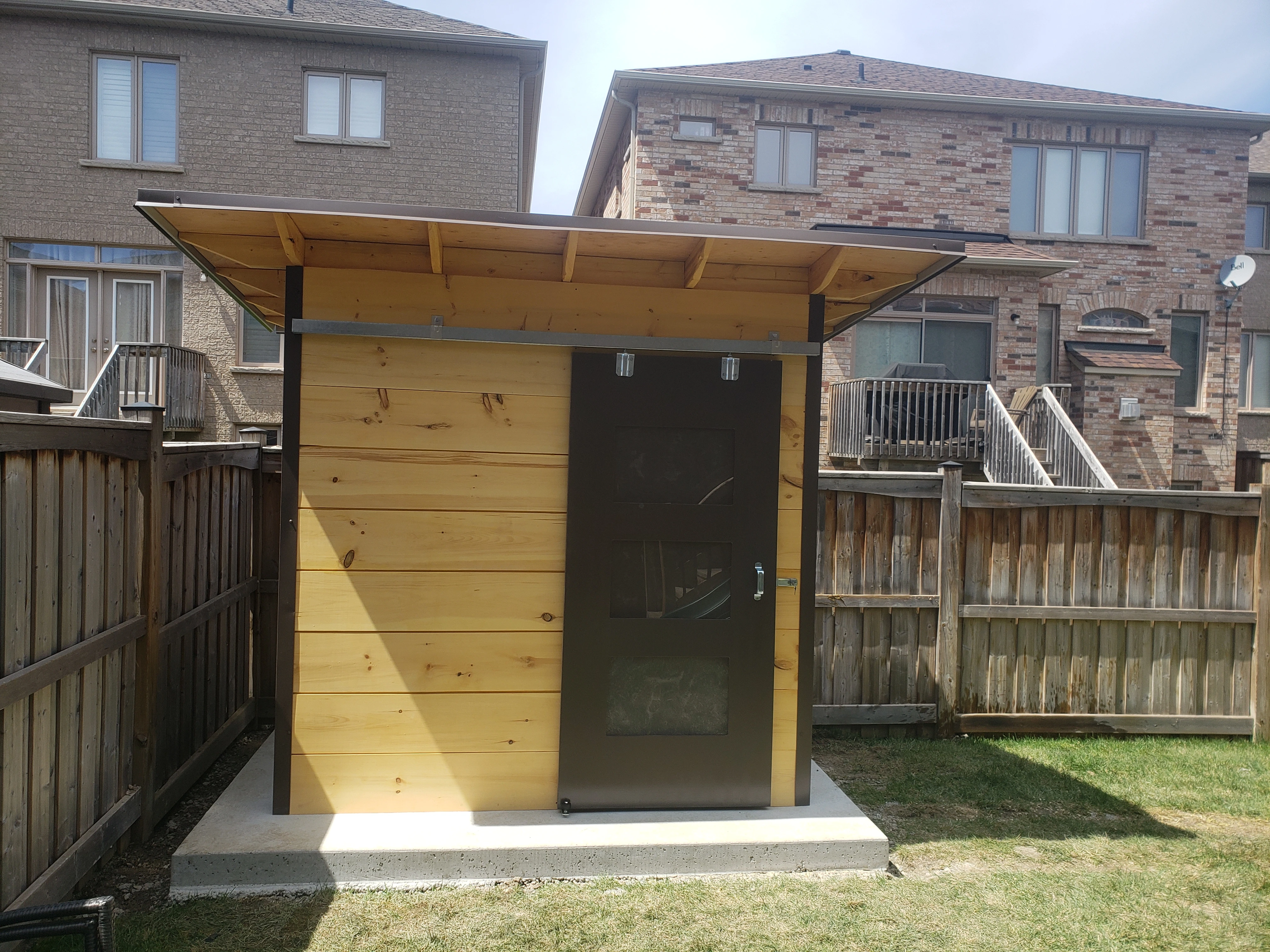 8x6 Essential Garden Shed with Metro Brown corrugated steel siding, horizontal pine shiplap front wall, and extended roof overhang with sliding barn door installed in Brampton Ontario by Backyard Escape Studios.