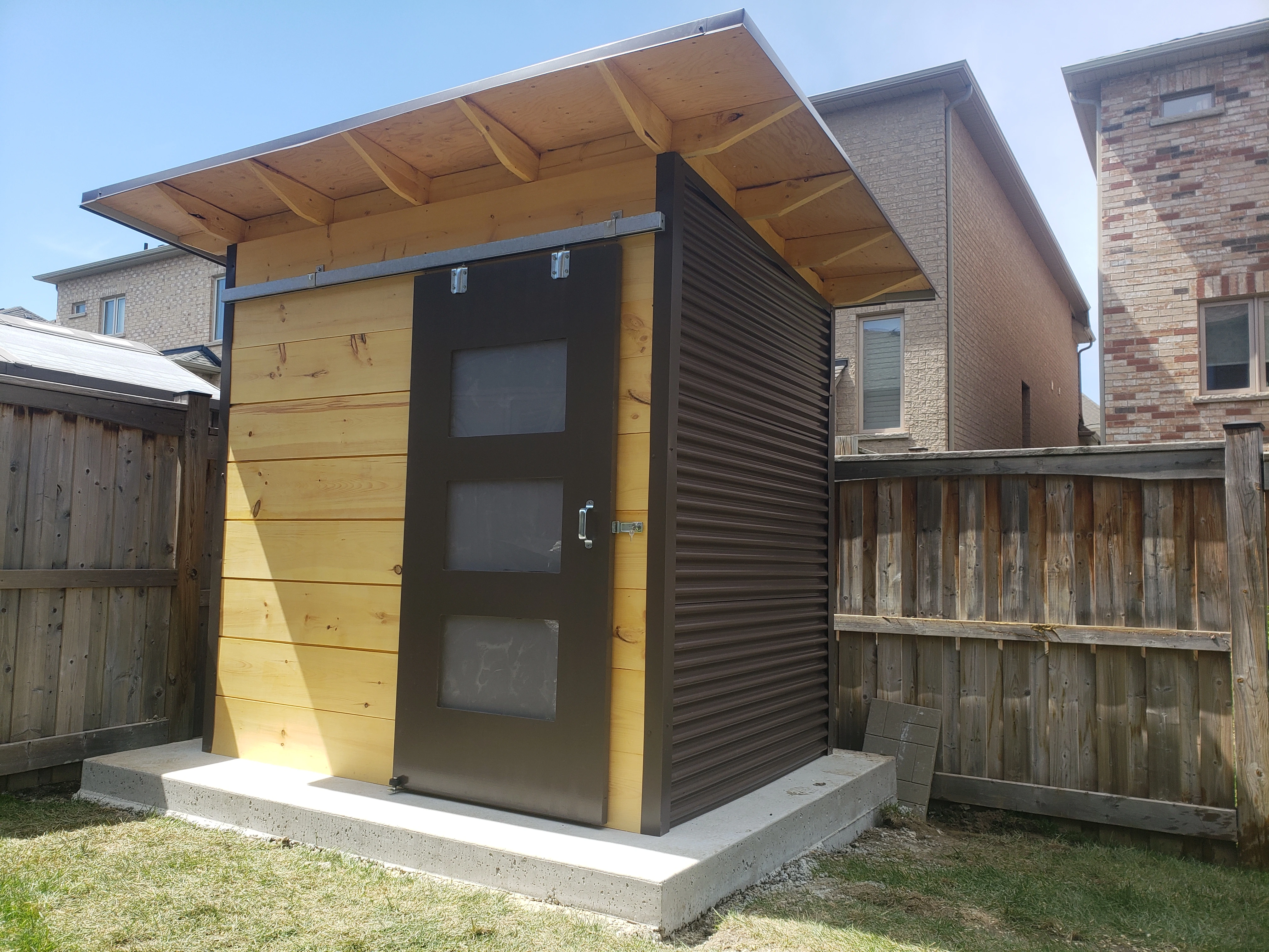 8x6 custom garden shed in Brampton Ontario showing sliding Metro Brown barn door with frosted glass lites and extended clear-coated rafter overhang, installed by Backyard Escape Studios.