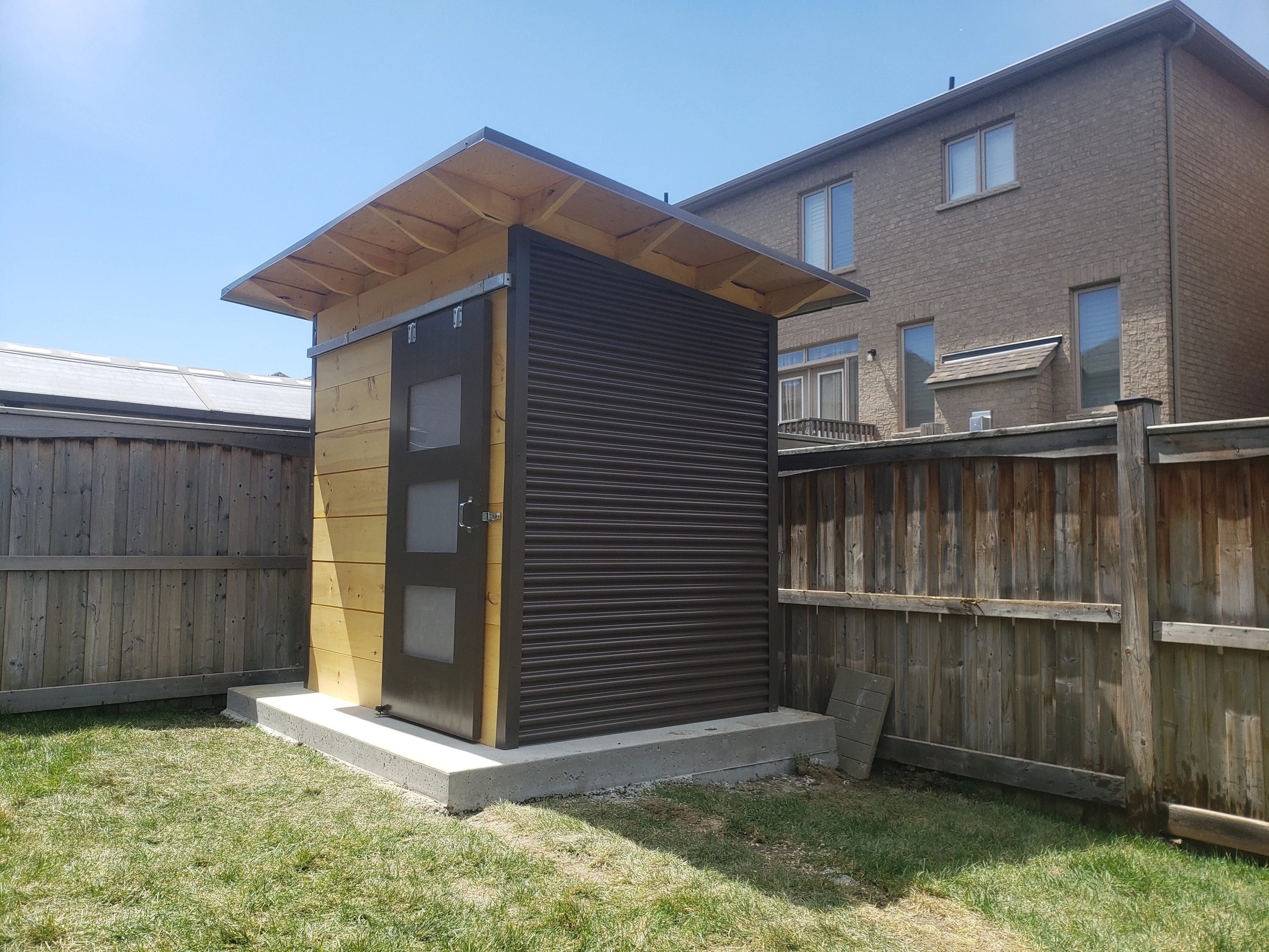 Front view of 8x6 Essential Garden Shed in Brampton Ontario with natural pine shiplap cladding, Metro Brown sliding barn door with triple frosted glass lites, and exposed rafter overhang, installed by Backyard Escape Studios.