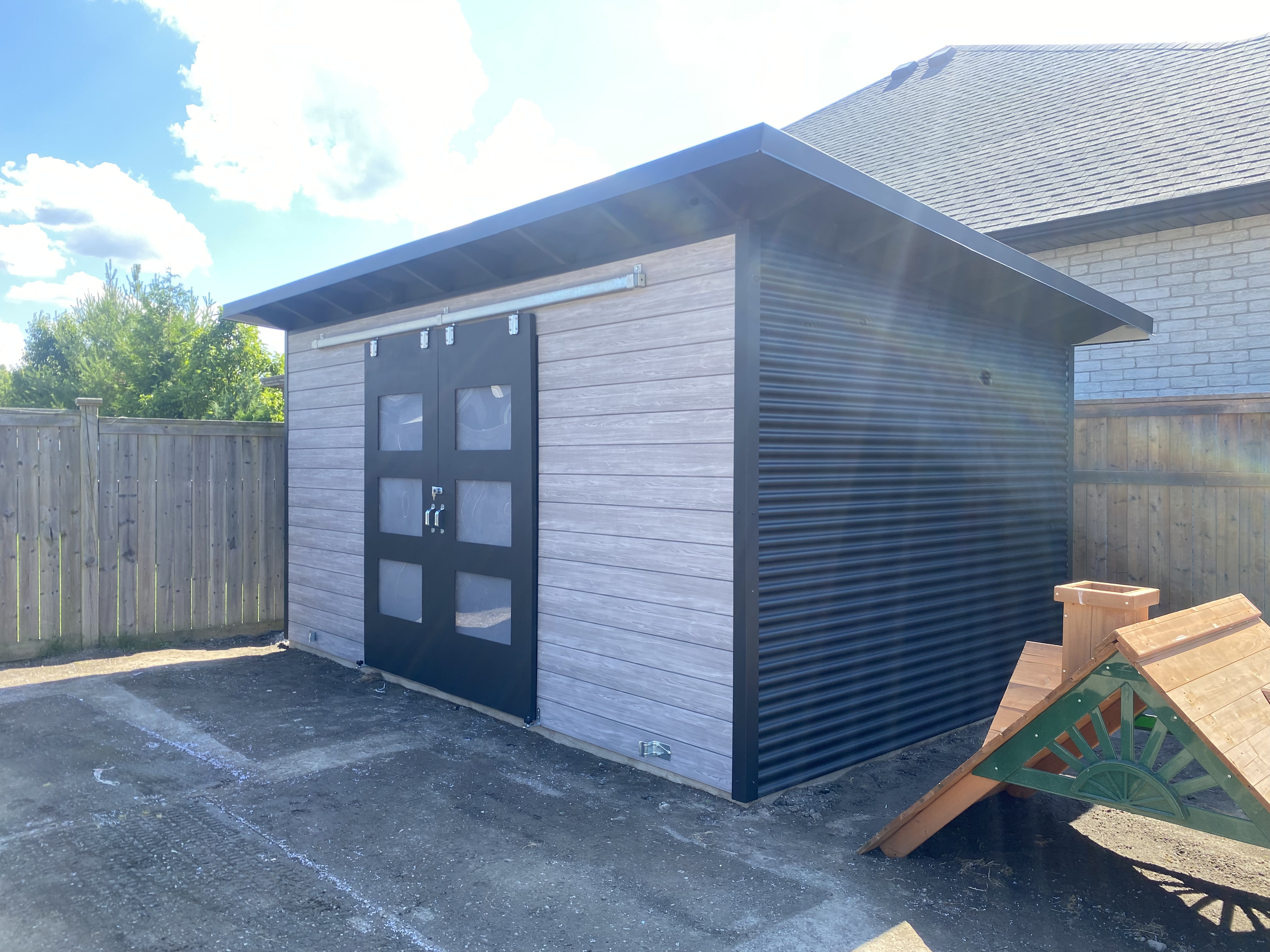 16x10 Storage Shed Essential Shed with Barnboard Grey Chamclad front, Jet Black corrugated steel sides, and double black sliding barn doors with frosted glass, installed in Sarnia, Ontario by Backyard Escape Studios.