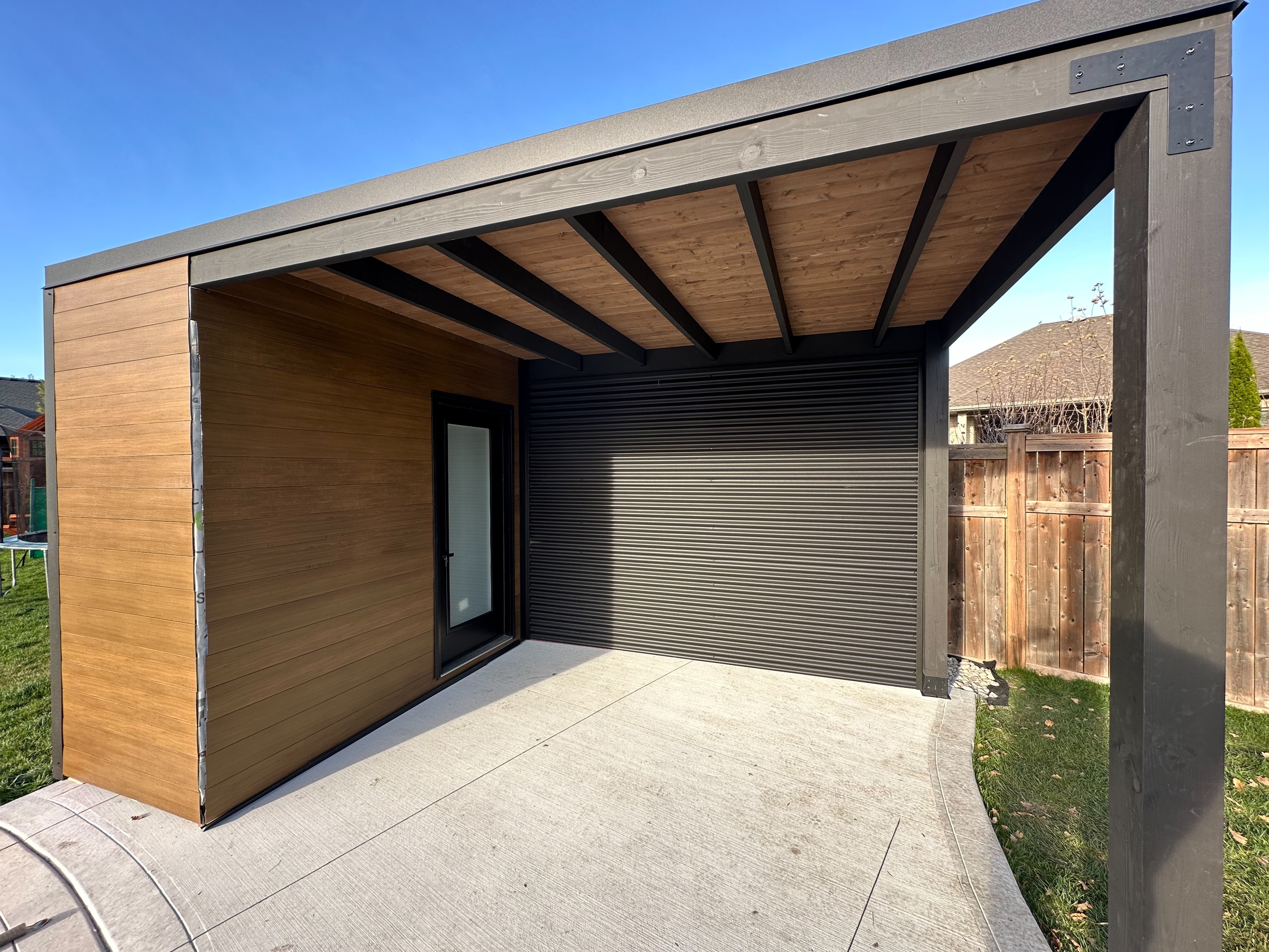16x9 Pool Cabana with jet black corrugated steel, teak shiplap siding, and a frosted glass change room door, installed beside an in-ground pool in Sarnia, Ontario by Backyard Escape Studios.