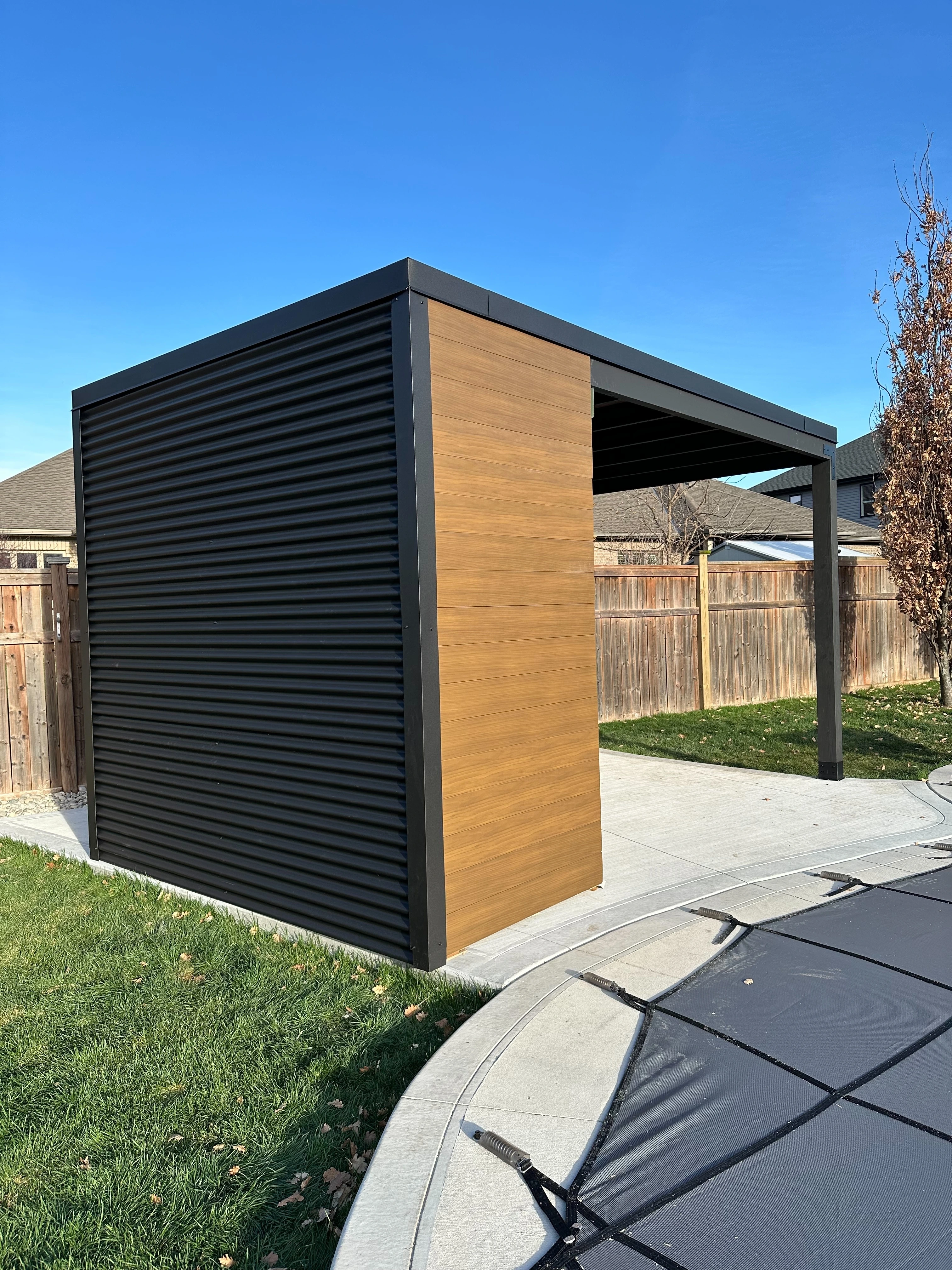 Side profile of a 16x9 Pool Cabana with jet black steel and teak shiplap, viewed from the pool deck in Sarnia, Ontario.