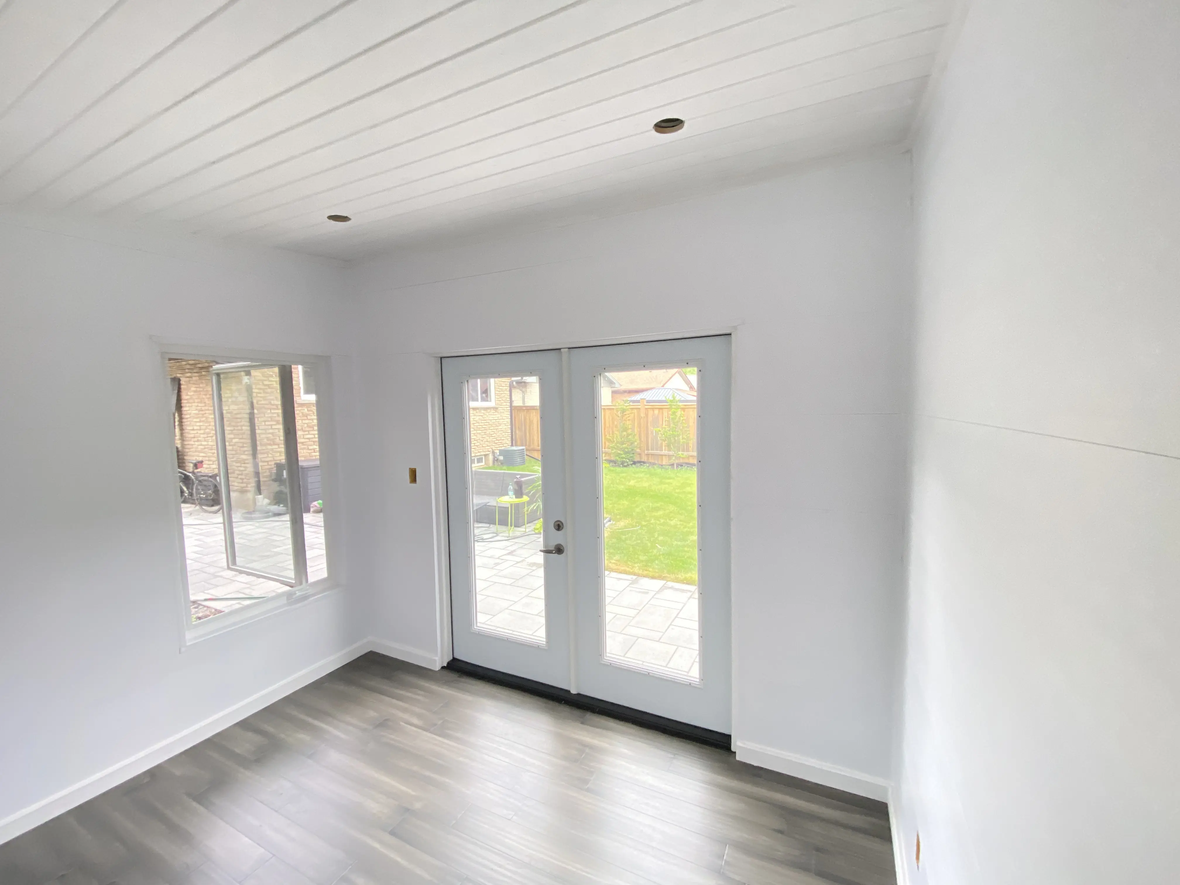 Alt: Finished interior of an 11x9 Backyard Office Studio in St. Catharines, Ontario with white Trus Core walls, pine T+G ceiling, grey vinyl plank flooring, and clear glass French doors.