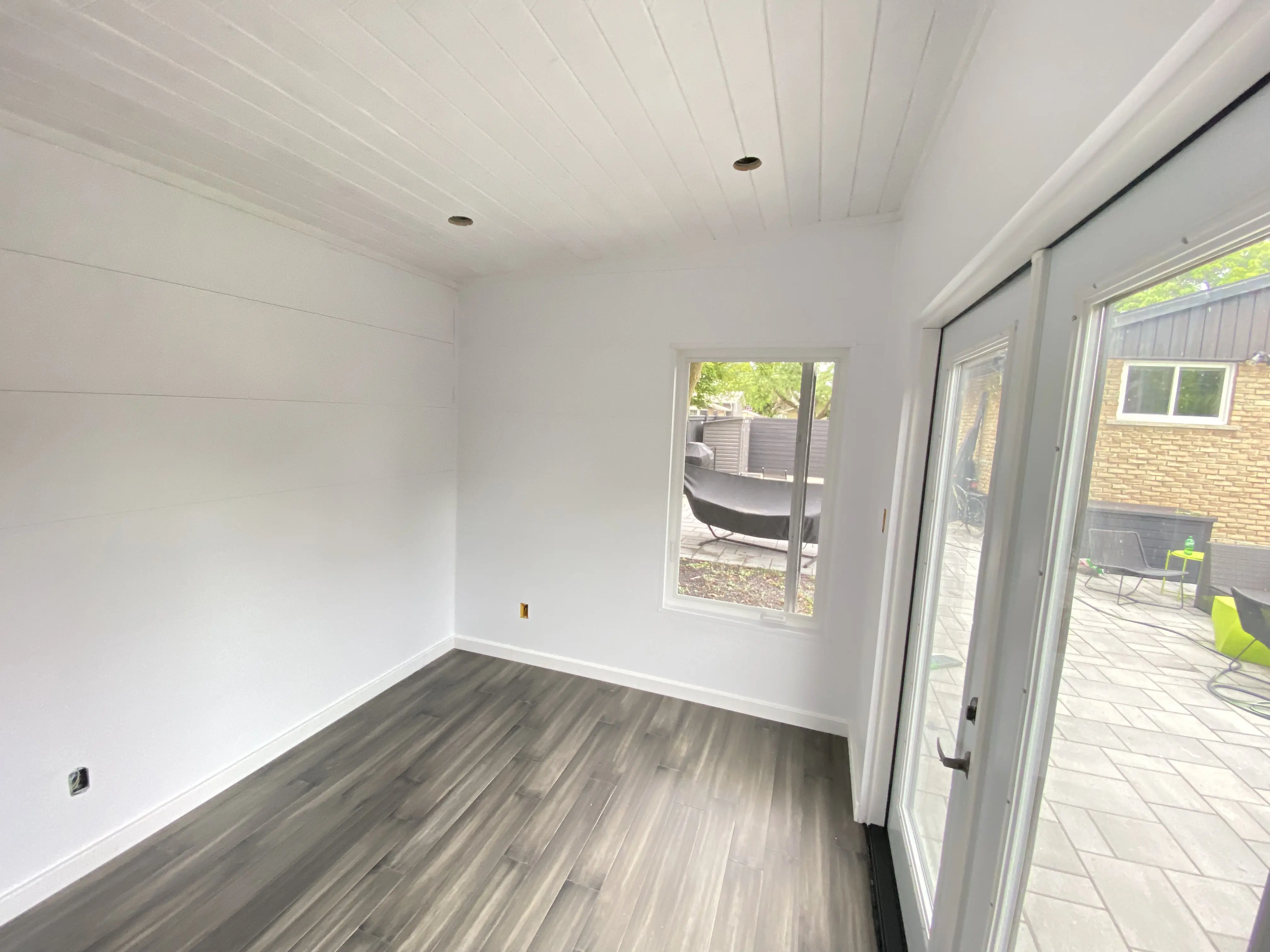 Alt: Interior corner of a Backyard Office Studio in St. Catharines, Ontario showing 36x60 casement window, white walls, recessed lighting, and grey vinyl plank flooring.
