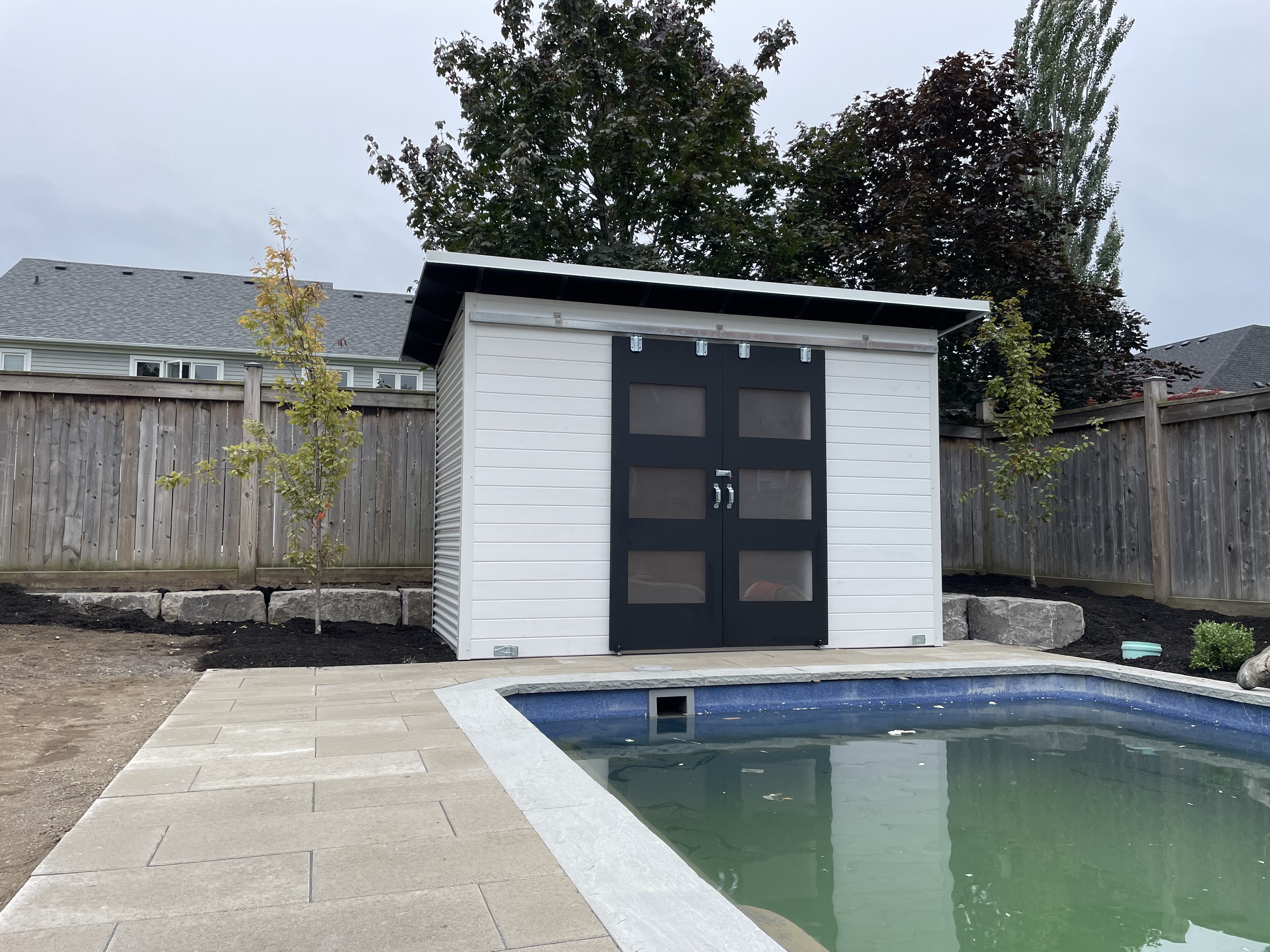 12x6 Pool Shed in White Satin pine T+G and Cambridge White corrugated steel with Black Beauty double sliding barn doors, Essential Shed installed on a pool deck in Whitby Ontario by Backyard Escape Studios.