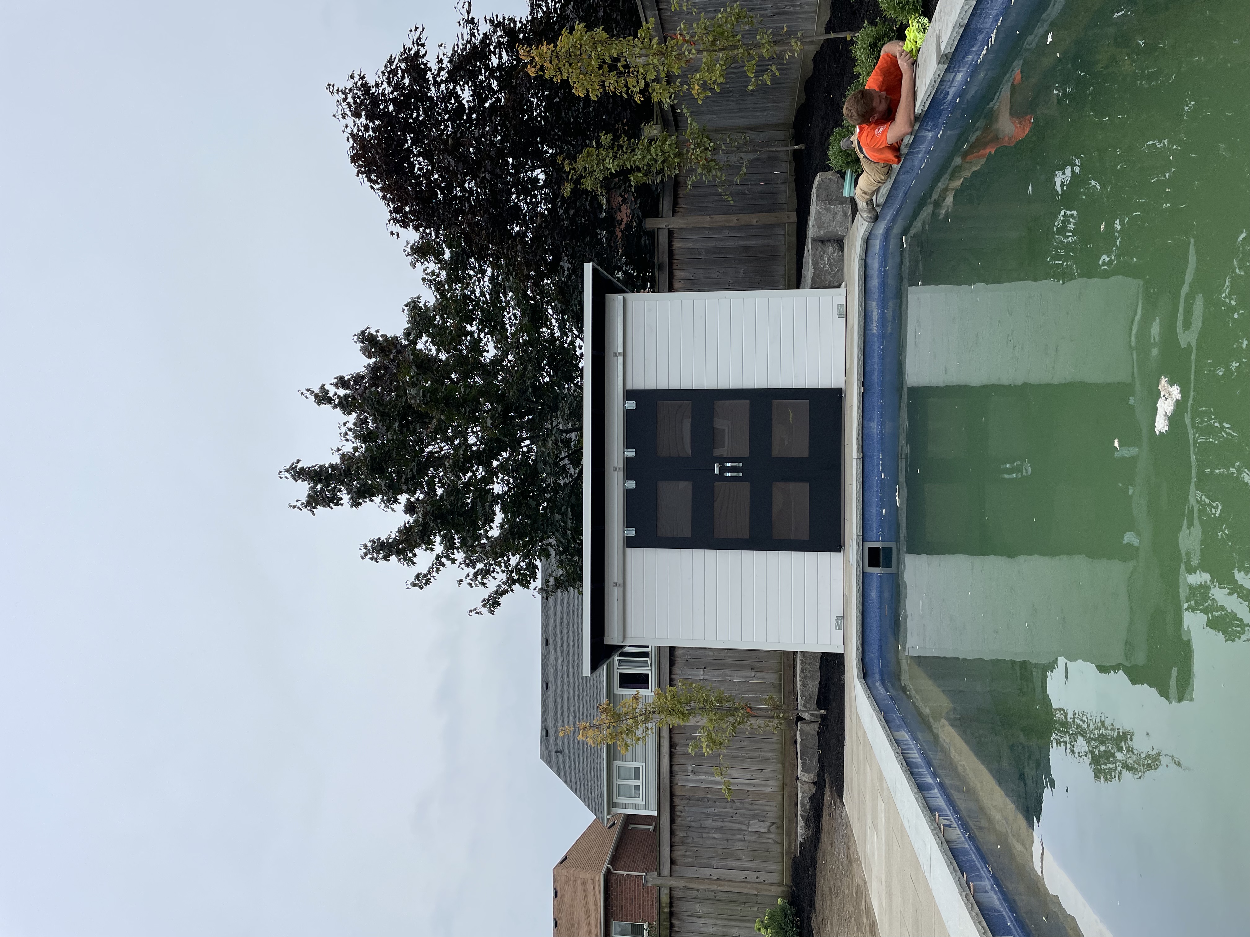 12x6 Pool Shed with Black Beauty double sliding barn doors and White Satin pine T+G front viewed across the inground pool in Whitby Ontario, Essential Shed by Backyard Escape Studios.