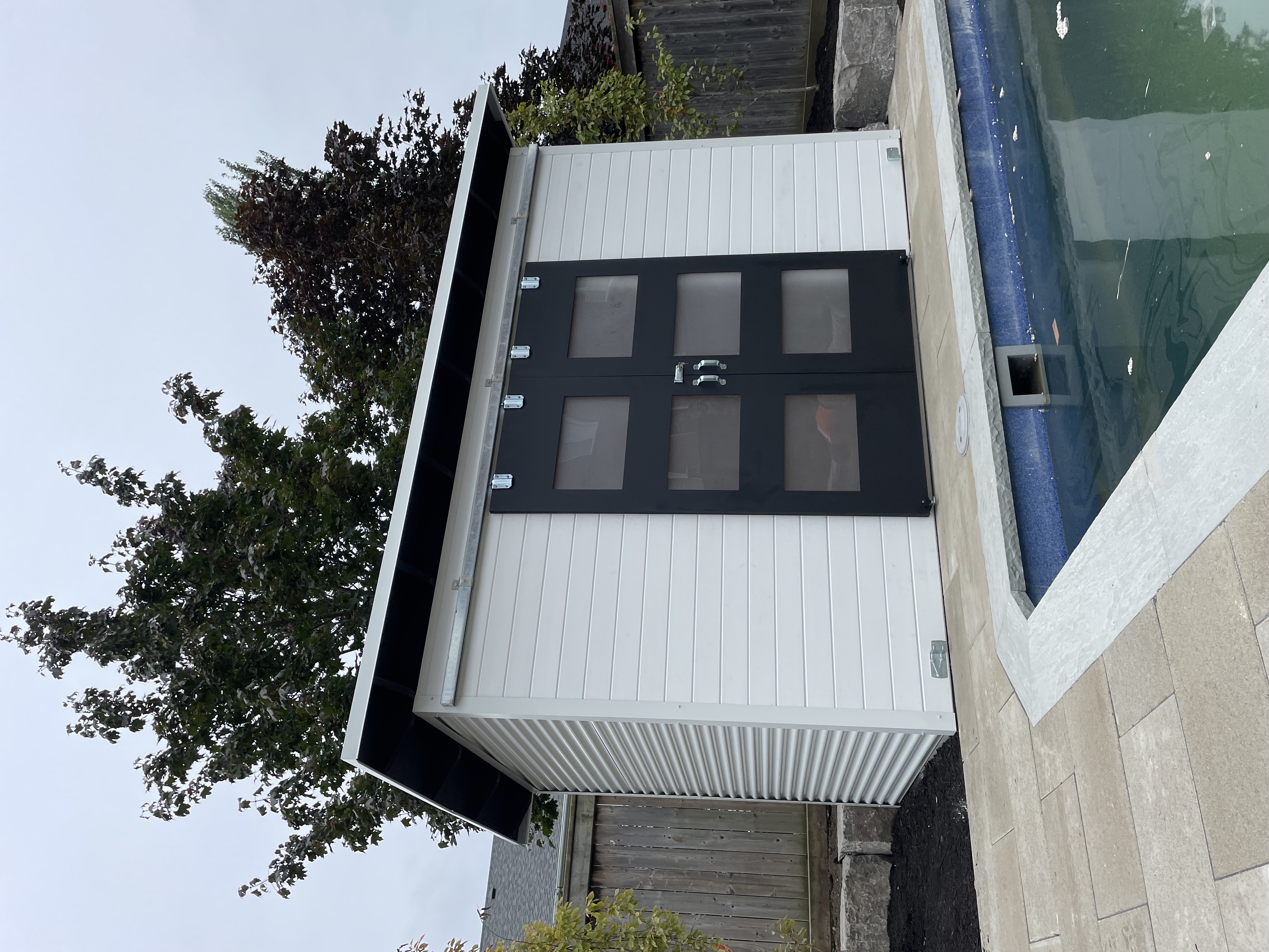 12x6 Pool Shed showing Cambridge White corrugated steel side wall and Black Beauty sliding barn doors, Essential Shed installed on a stone pool deck in Whitby Ontario by Backyard Escape Studios.