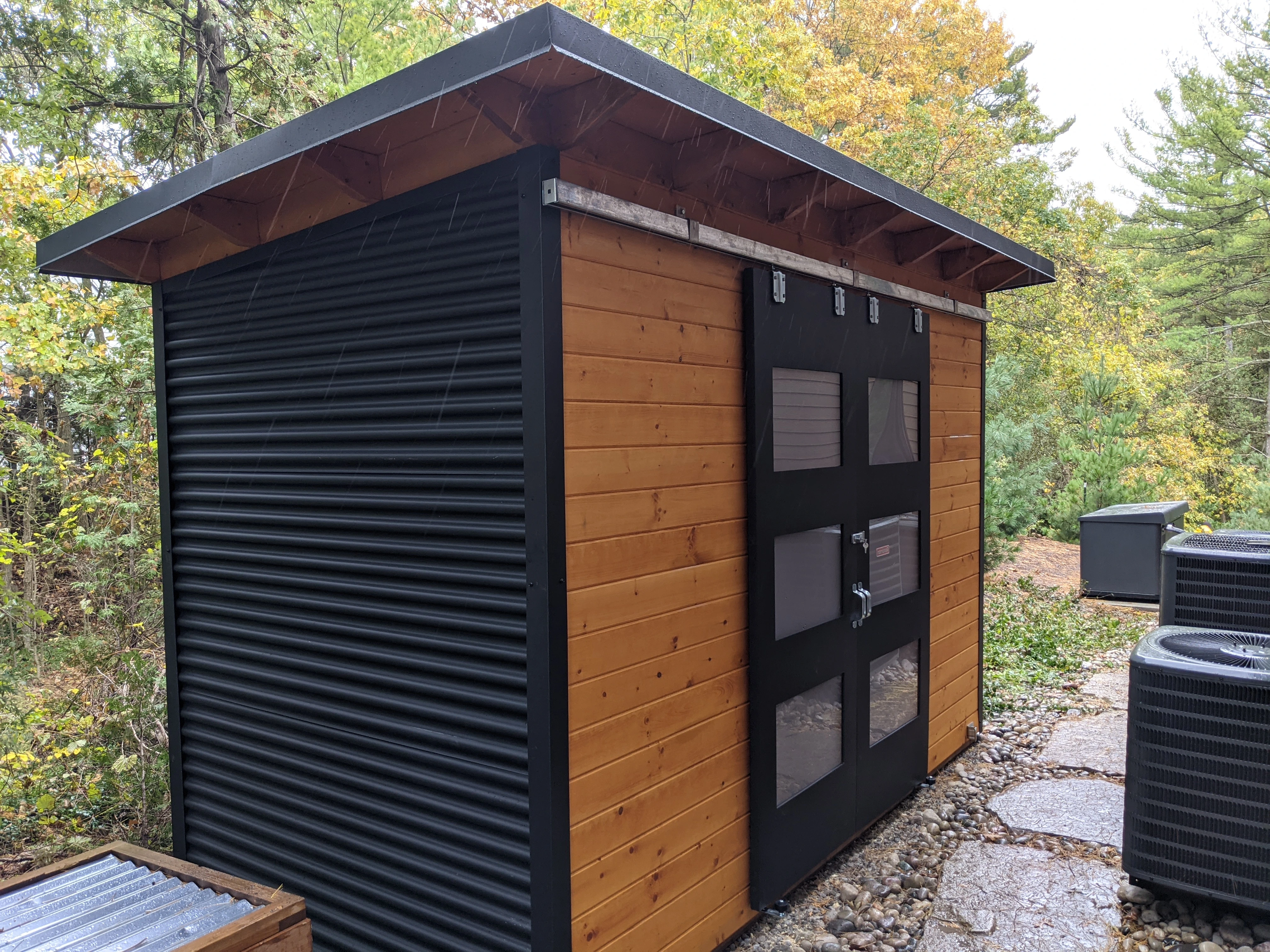 12x6 Essential Storage Shed with Jet Black corrugated steel siding, cedar-stained pine T+G front, and double sliding barn doors with frosted Lexan windows, installed on a lakeside Huron Drive property in Grand Bend, Ontario by Backyard Escape Studios.