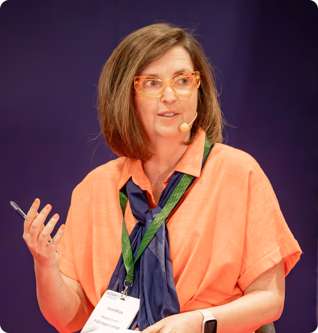 Woman with shoulder-length brown hair and orange glasses speaking, wearing an orange shirt, navy scarf, and green lanyard with a name tag.