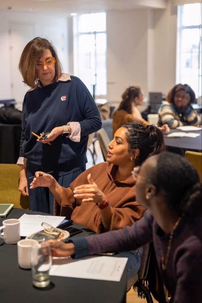 Woman in glasses standing and two seated women discussing papers at a workshop table.