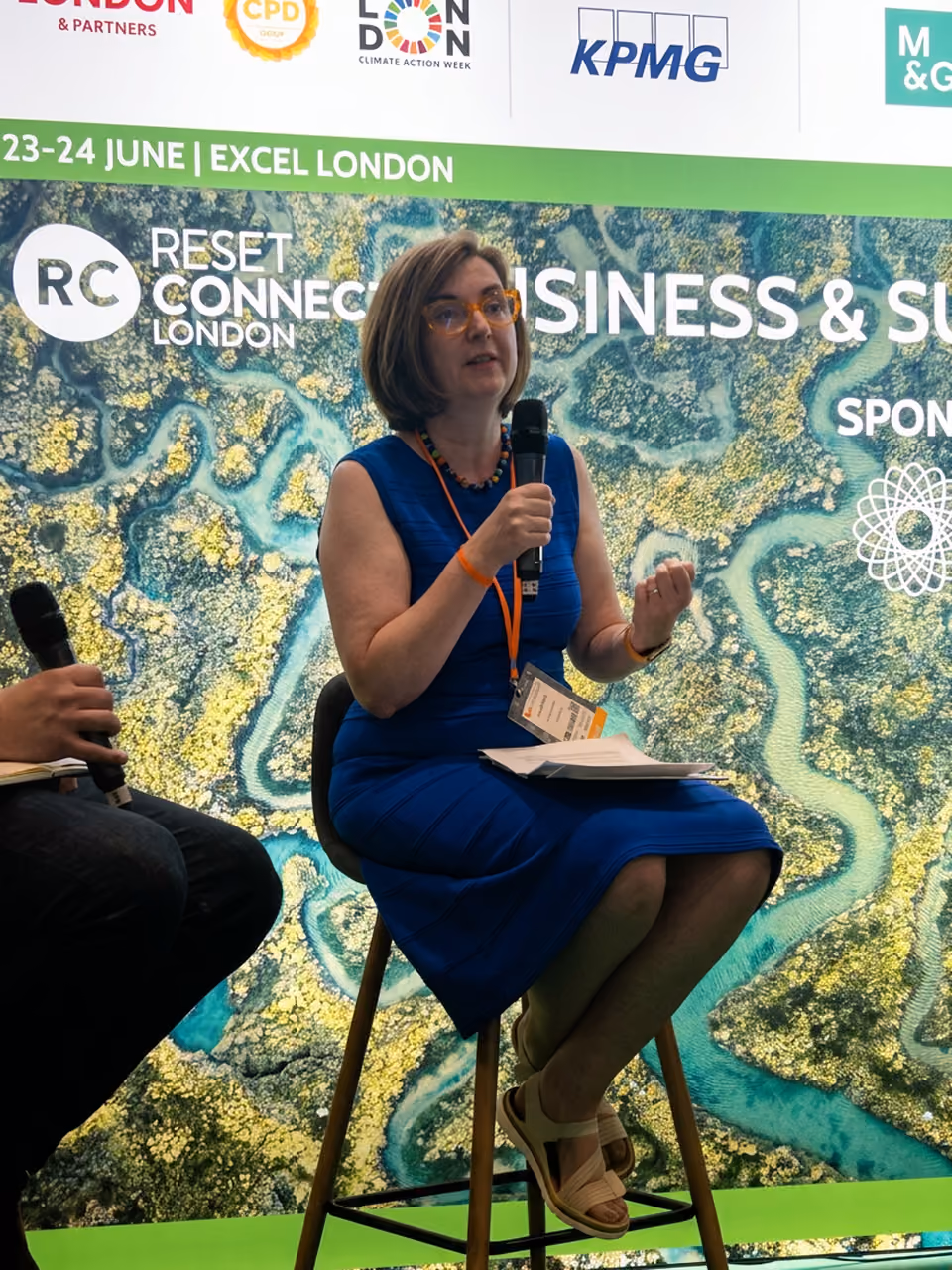 Woman in blue dress sitting on stool holding microphone and papers at a Reset Connect London event.