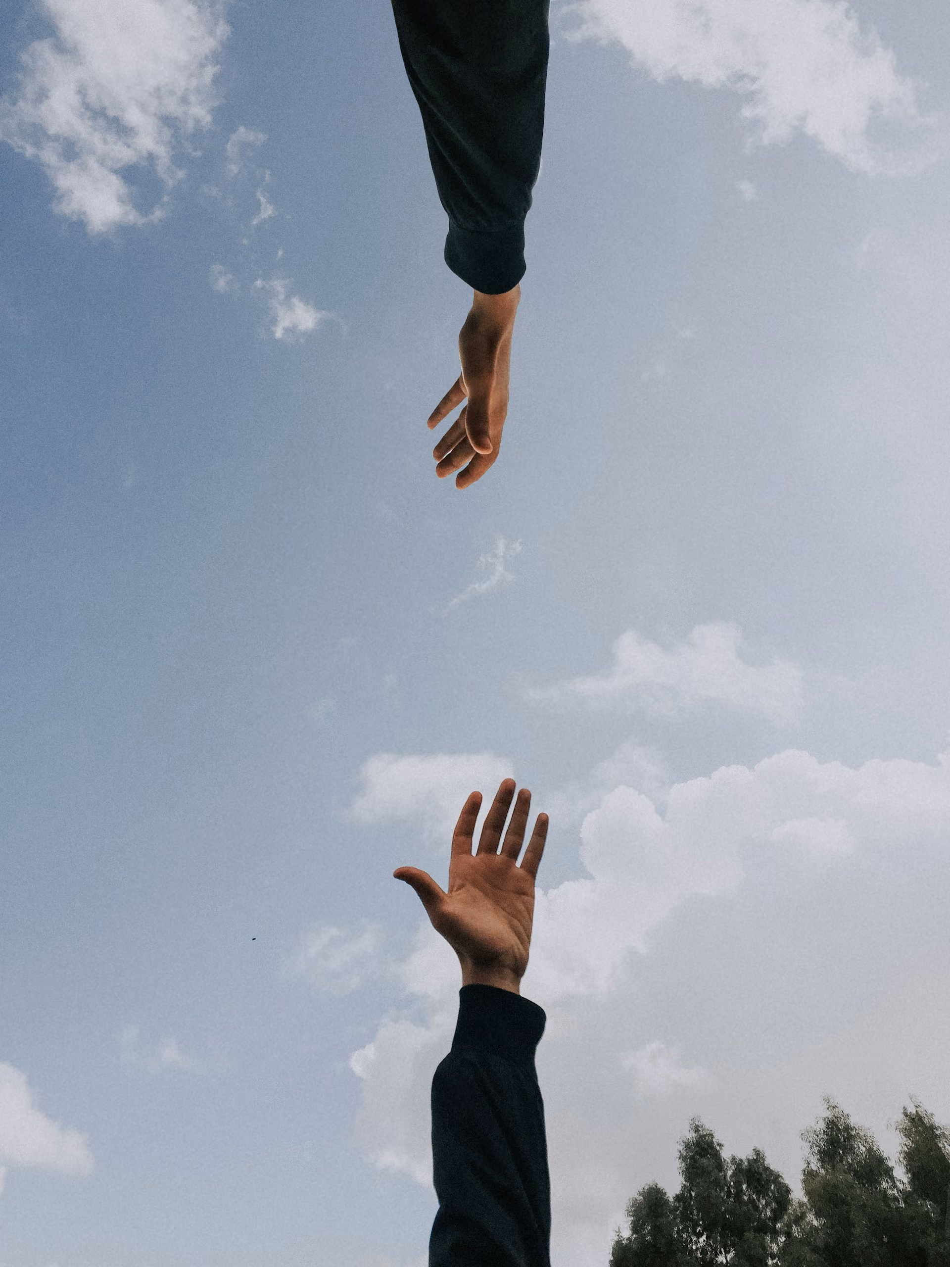 Two hands reaching toward each other against a blue sky with clouds and some tree tops.