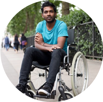 A male with black hair in a wheelchair smiling with his arms resting above his legs crossed.