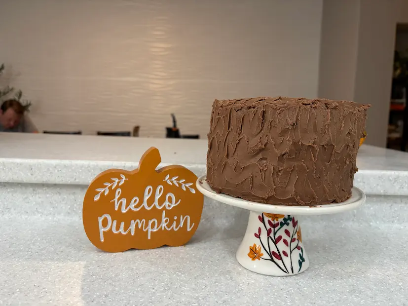 Chocolate-frosted pumpkin chocolate chip cake on a cake stand beside a "hello pumpkin" sign