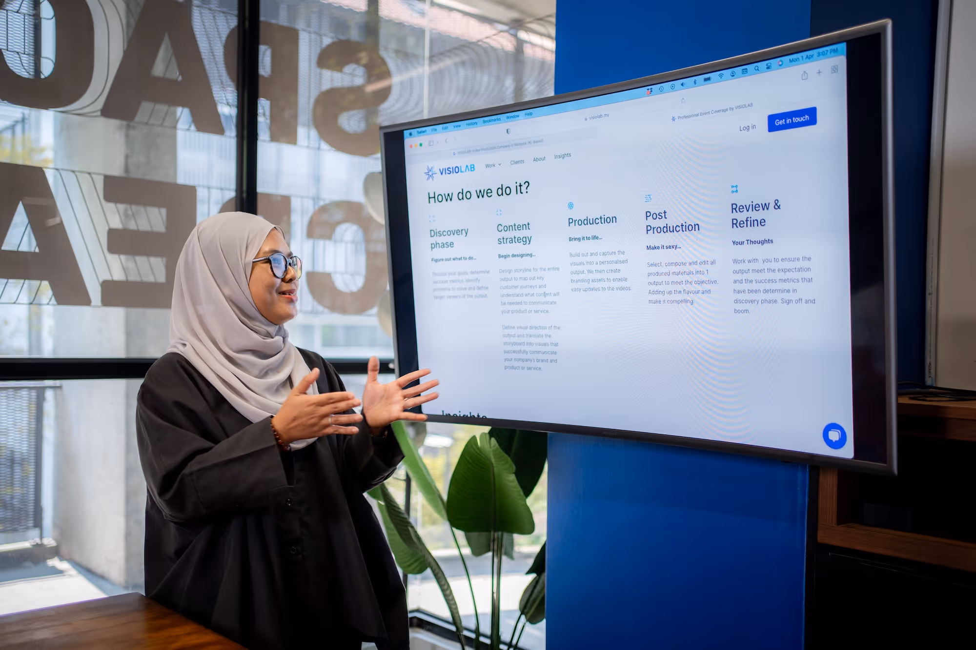 Woman in hijab and glasses gesturing while presenting a content strategy and production process on a curved screen.