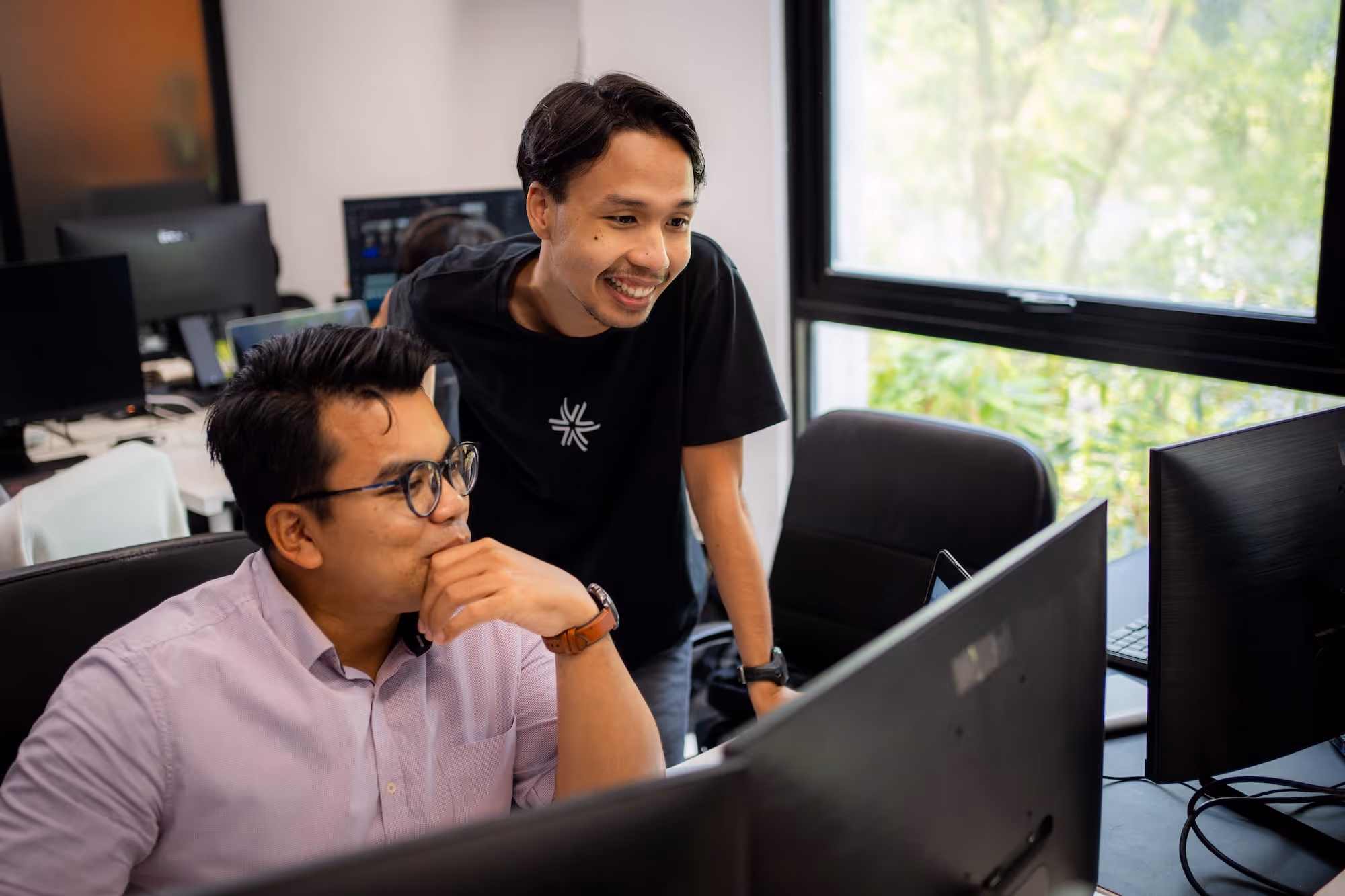 Two men collaborating at computer monitors in a modern office with a large window.