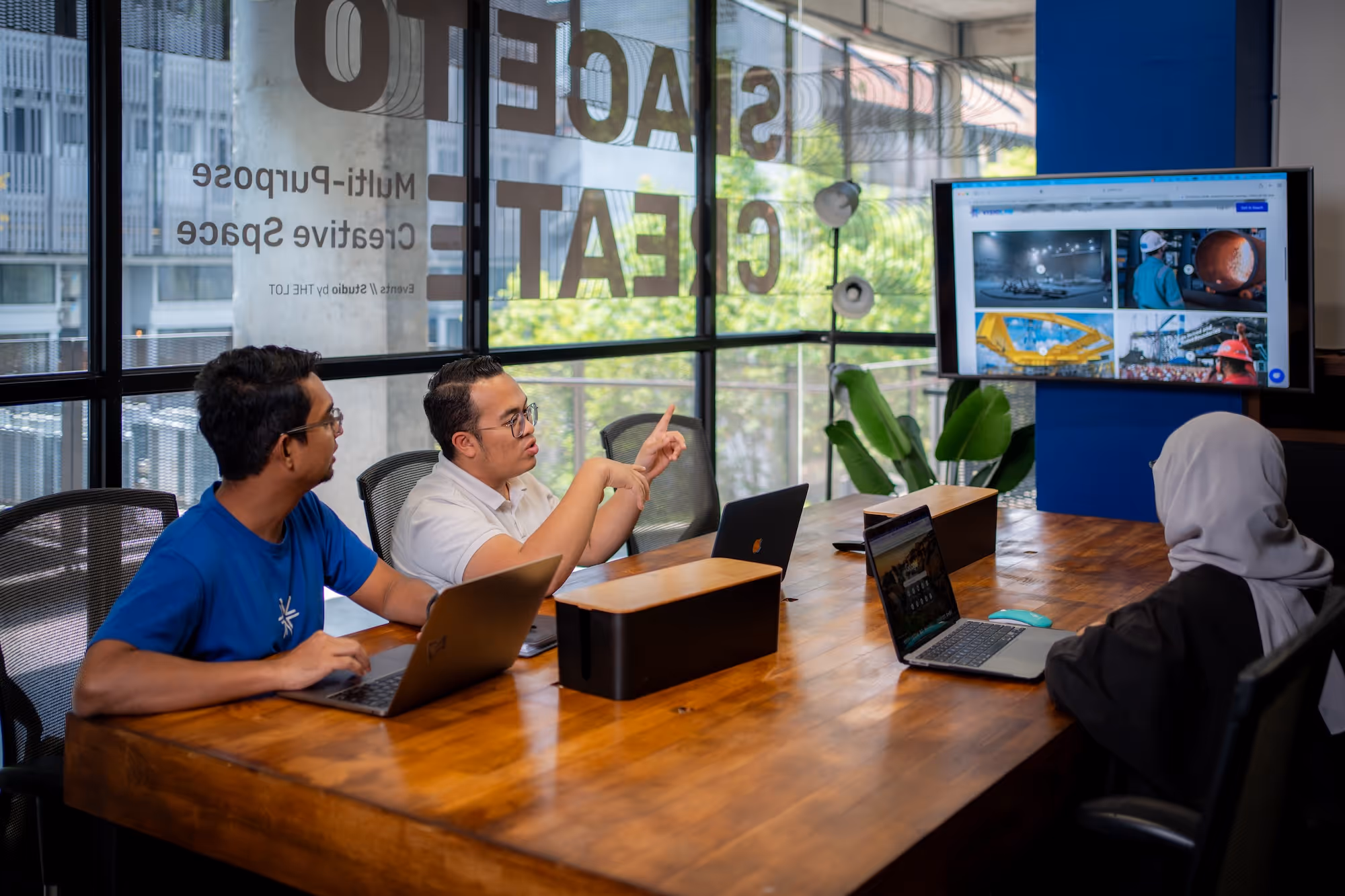 Three people in a meeting room working on laptops while discussing and looking at a screen with industrial images.