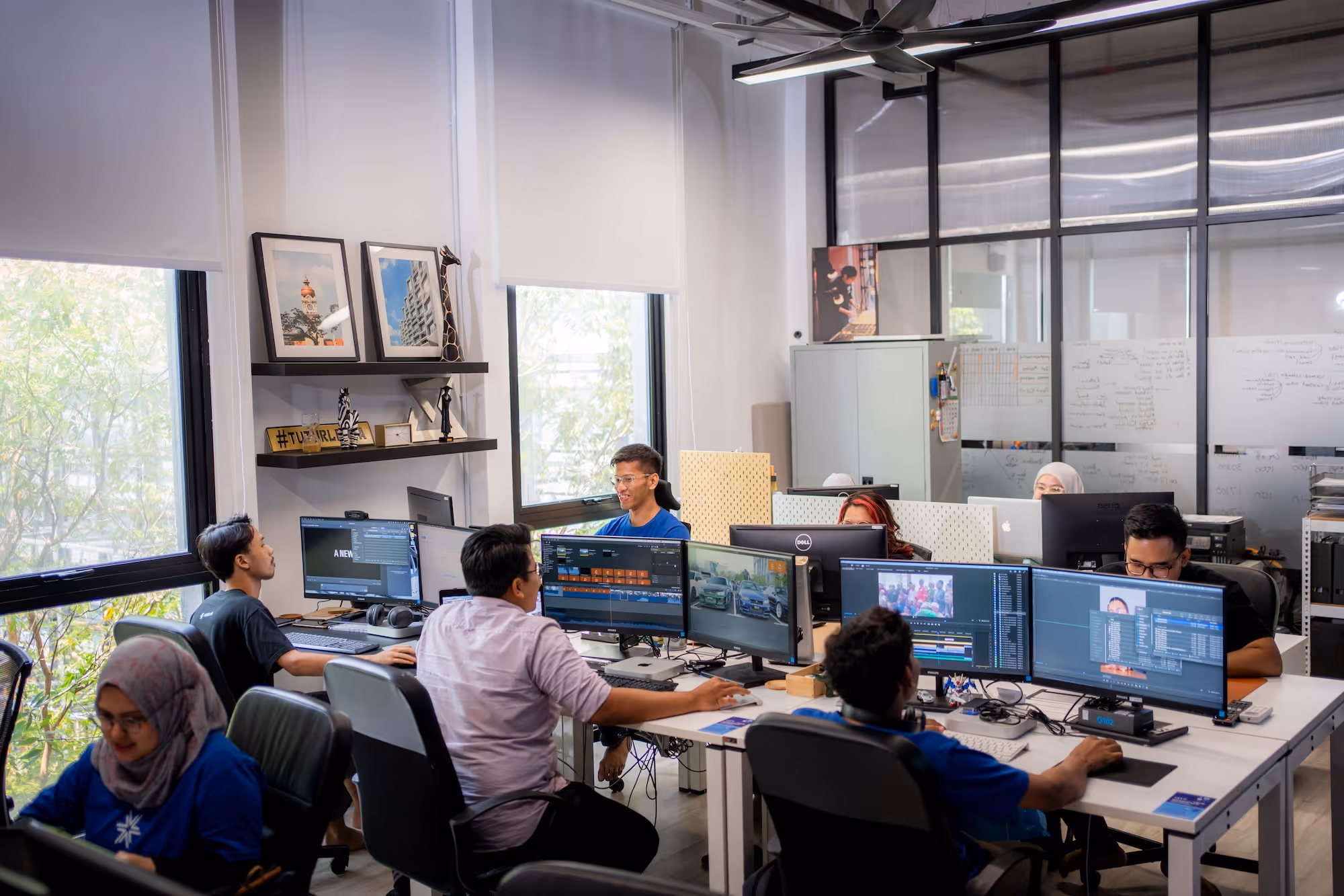 Office space with seven people working on computers with multiple monitors and video editing software.