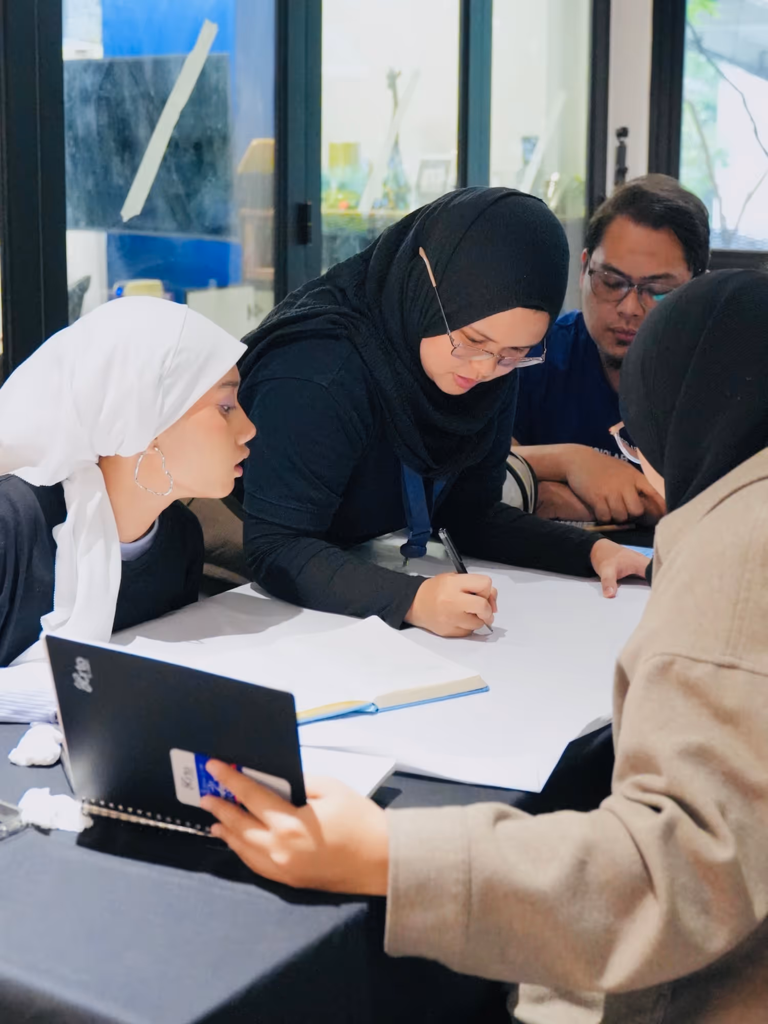 Four people in hijabs and glasses collaborate around a table with notebooks and a large white paper.