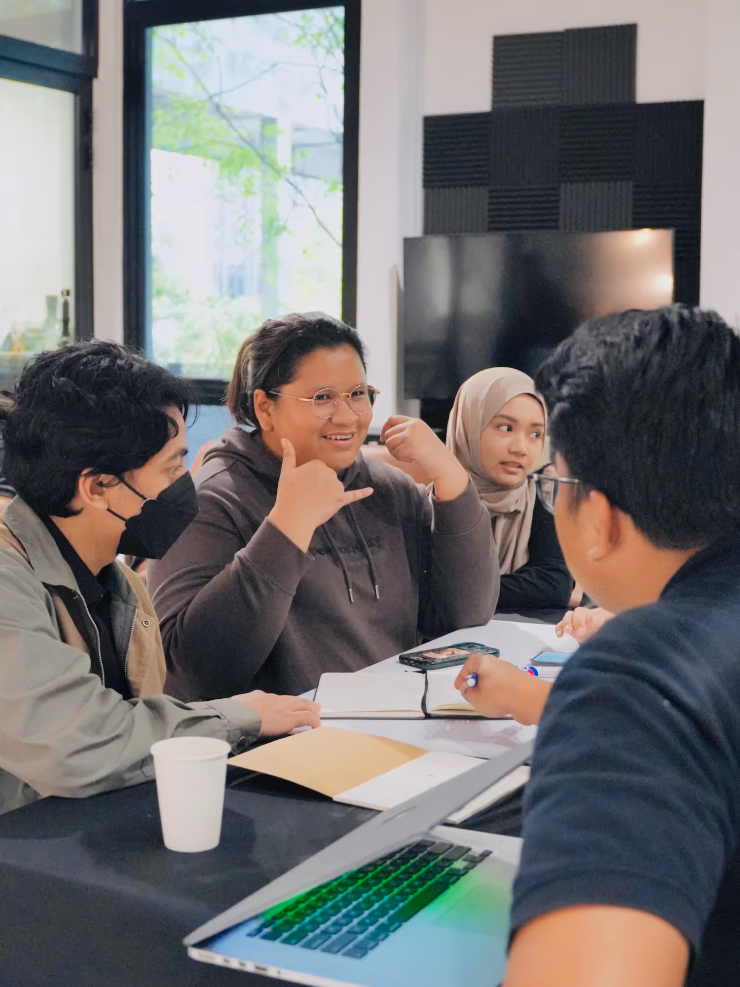 Four young adults gathered around a table with laptops and notebooks, engaged in discussion in a bright room.