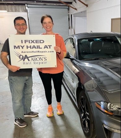 Couple with their gray Ford Mustang holding an I fixed my hail at Aaron's sign