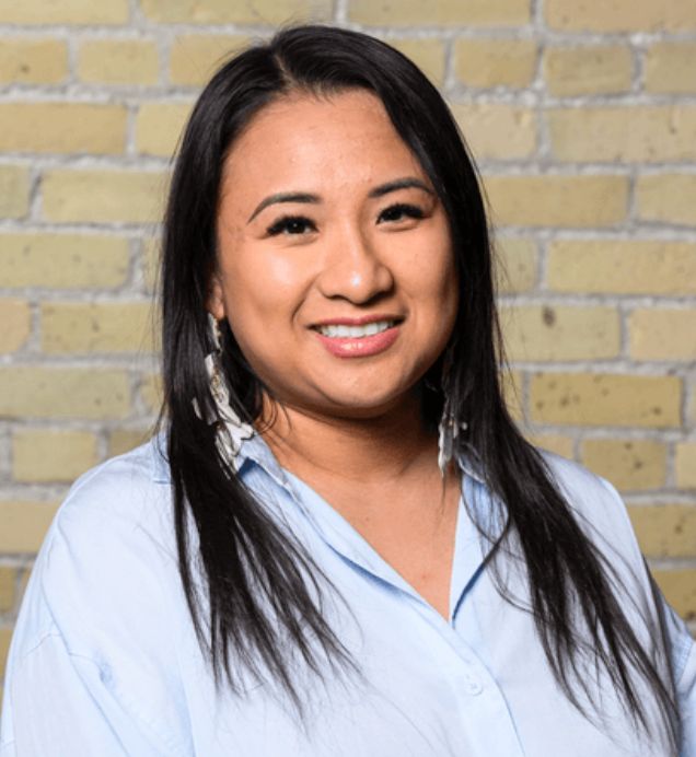Liz Cron, Advocacy & Stakeholder Relations — Smiling person with long dark hair wearing a light blue button-up shirt and beaded earrings, against a brick wall