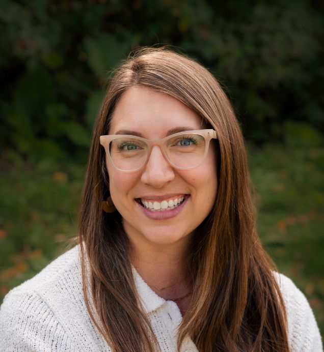 Rose Regier, Complex Engagement & Operations — Smiling person with long brown hair and glasses wearing a white sweater, outdoors with greenery behind