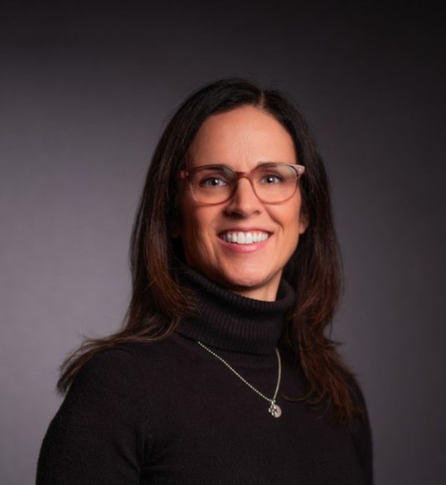 Andrea Aiello, Sector Development & Transformation — Smiling person with long dark hair and glasses wearing a black turtleneck and pendant necklace, against a dark background