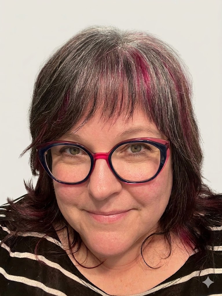 A person with shoulder-length dark hair streaked with magenta and gray highlights, wearing round red-and-blue eyeglasses and a black-and-white striped top. The person is looking at the camera with a slight smile against a plain light-colored background.