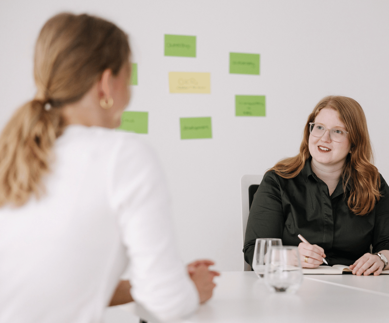 Im Hintergrund sind die Oberkörper und Hände von zwei Männern zu sehen, die sich an einem Tisch unterhalten. Im Vordergrund sind weiße Icons wie eine Zielscheibe und ein Balkendiagramm zu sehen