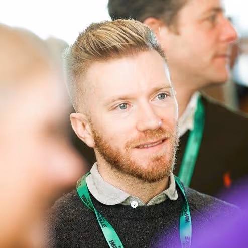 Luke Smith mit hellbraunem Haar und Bart, trägt ein grünes Schlüsselband und einen grauen Pullover und schaut leicht zur Seite.