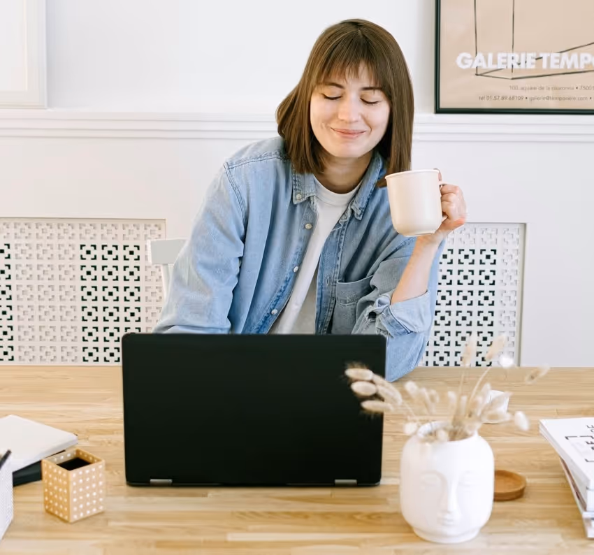 Young woman smiling with eyes closed, holding a coffee mug while sitting at a desk with a laptop.