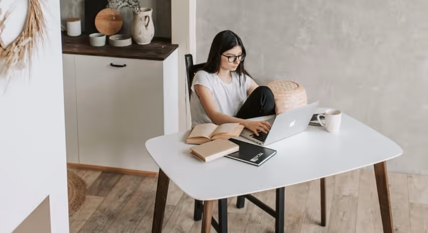 Young woman with glasses sitting at a white table using a laptop surrounded by books and a coffee mug in a modern room.
